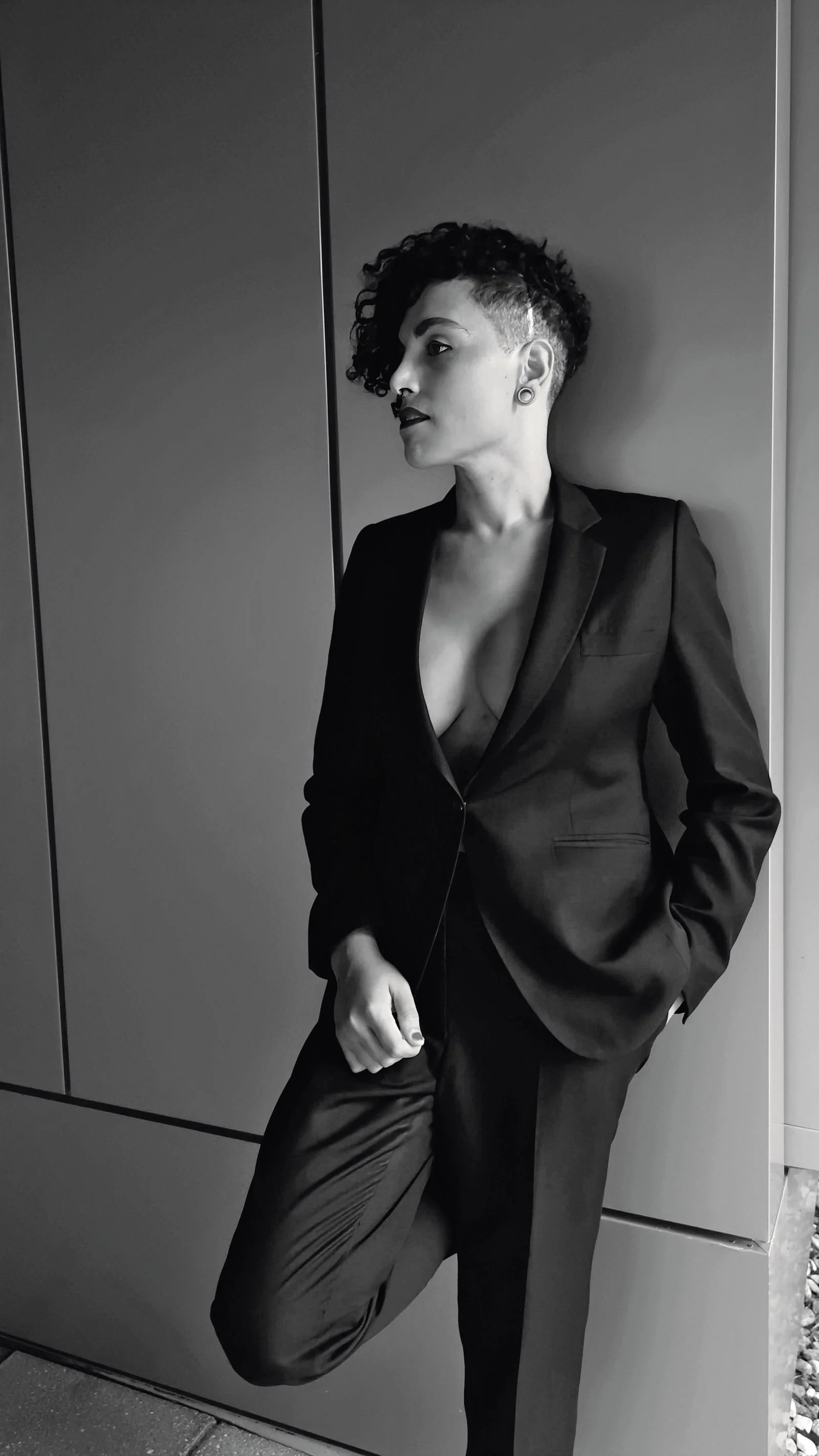 Black and white photo of a woman with a mohawk hairstyle, wearing a blazer and pants, standing against a wall.