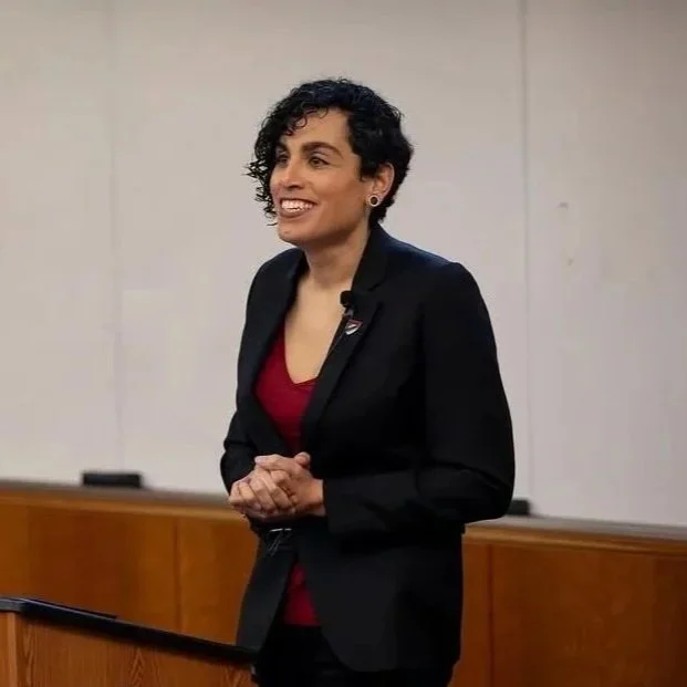 Alejandra Caraballo with short curly dark hair, wearing a black blazer and red shirt, smiling and speaking in front of a whiteboard in a classroom or lecture hall.