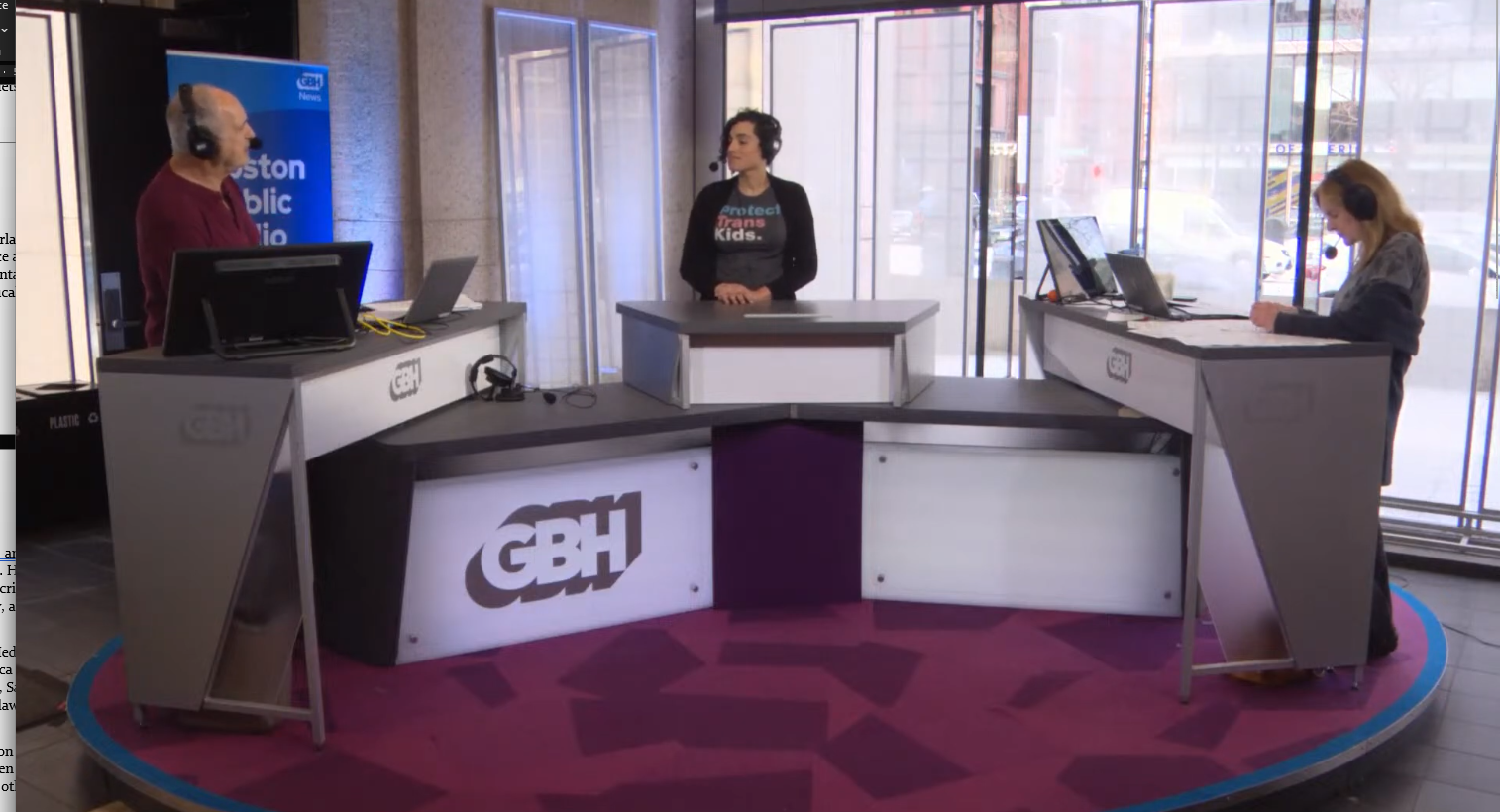Radio studio with three people at microphones and desks, GBH News logo, large windows in background.