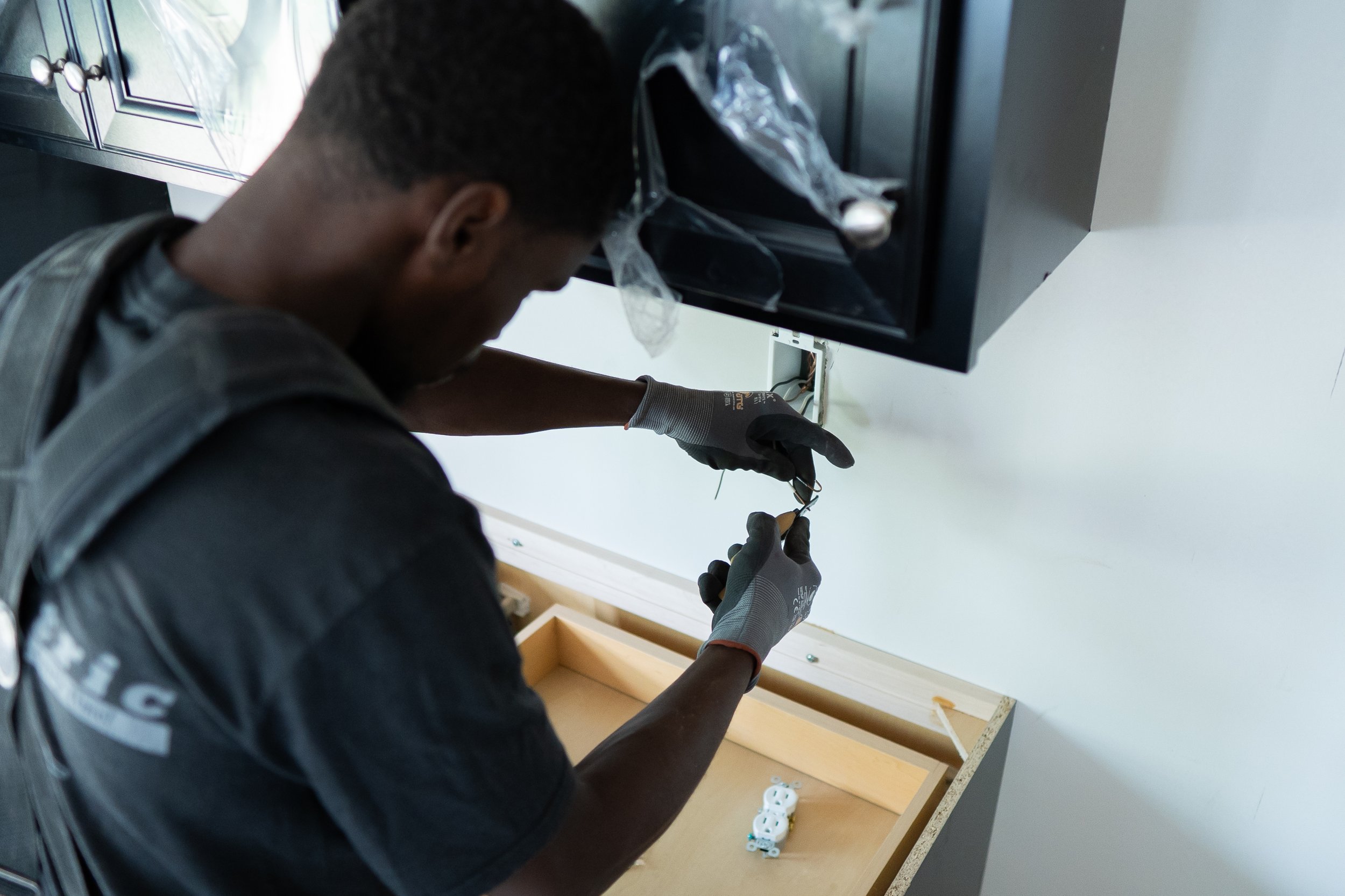 A man working on electrical wiring inside a wall behind a kitchen counter, wearing safety gloves.