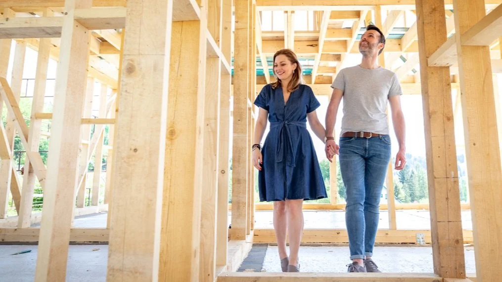 A smiling woman wearing a blue dress and a man in a grey t-shirt and jeans walking hand-in-hand through a house under construction with visible wooden framing.