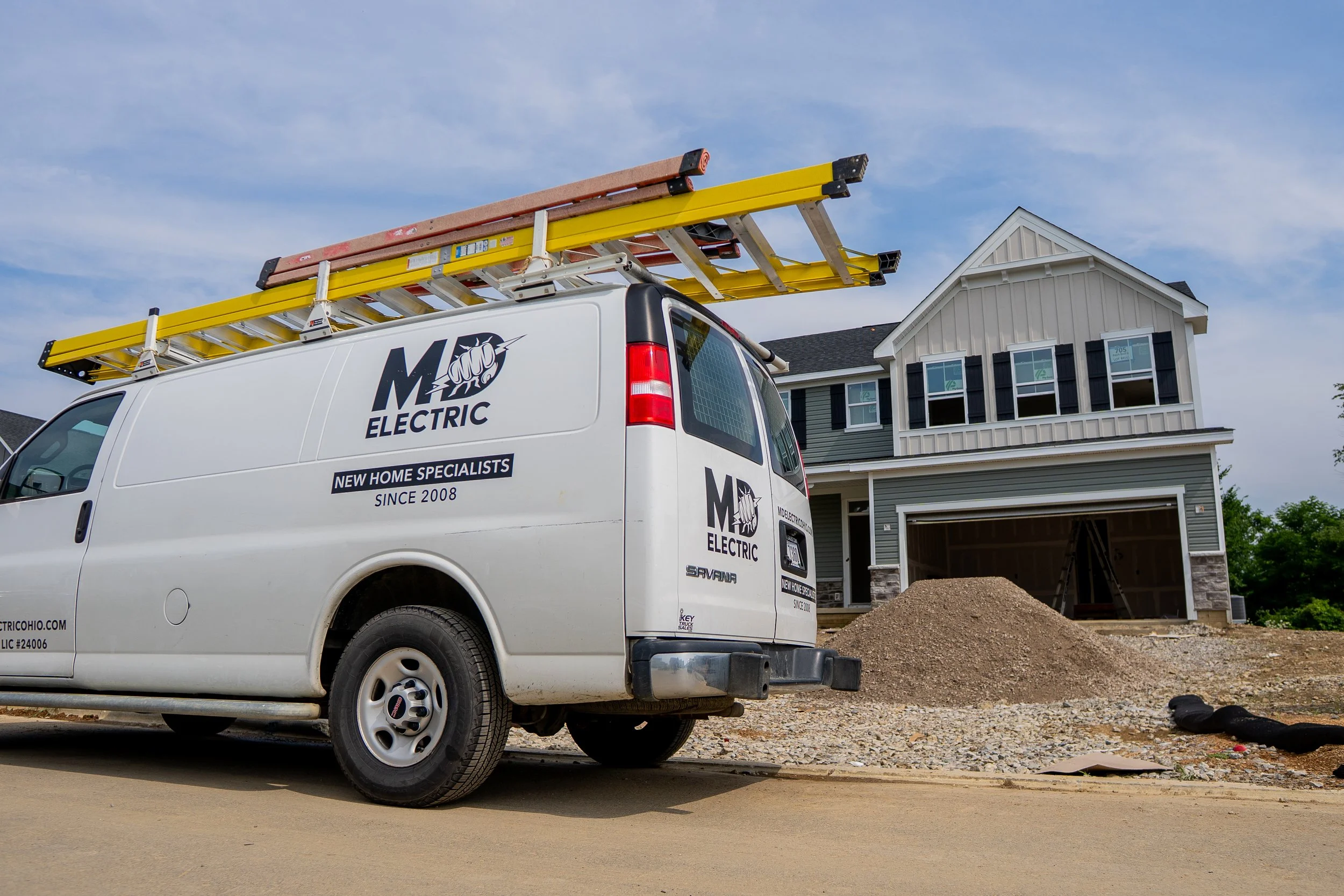 A white electric service van with a ladder on top, parked in front of a house under construction.