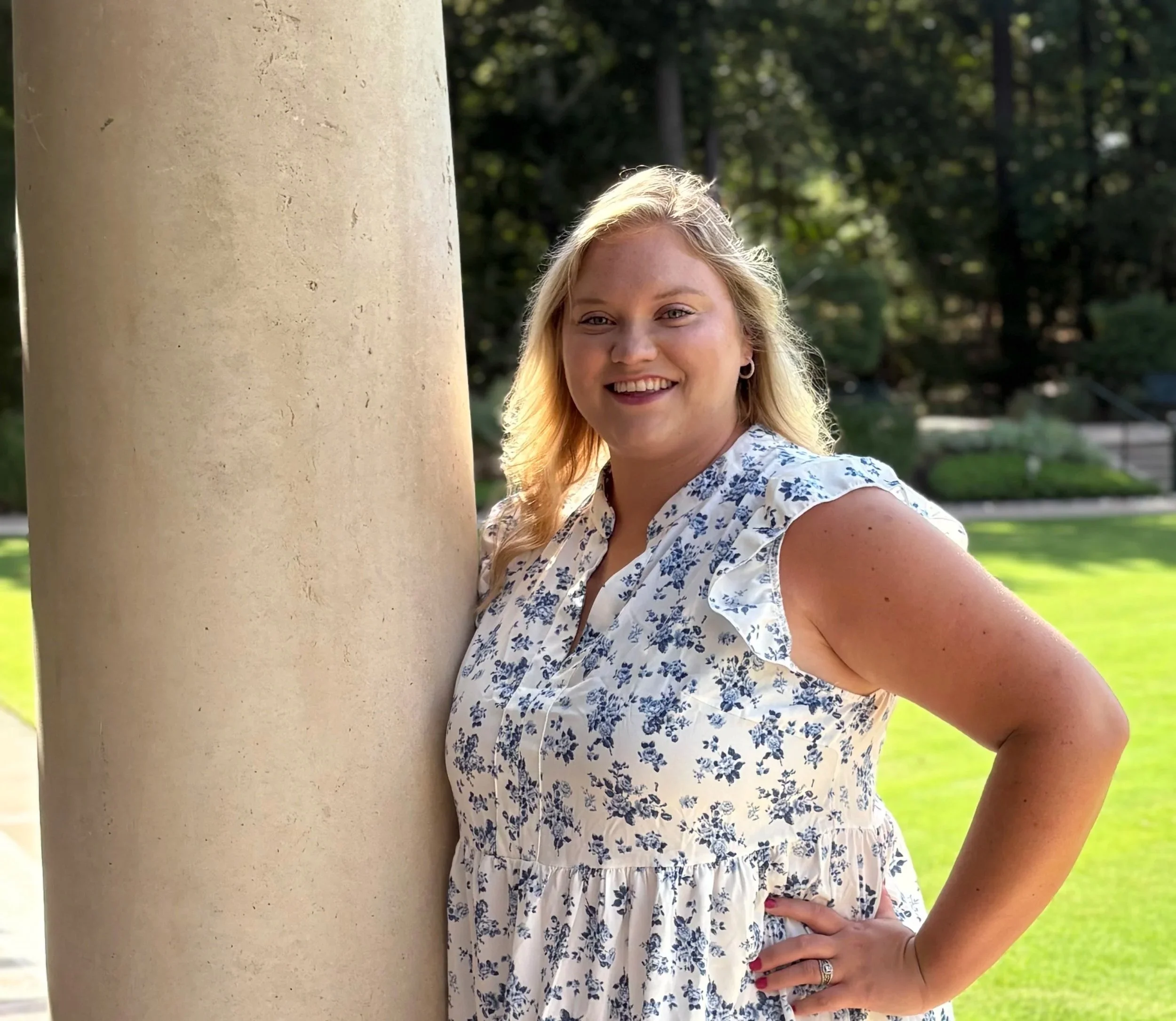 A woman with blonde hair in a blue and white floral dress leaning against a large beige column outdoors on a sunny day, smiling with one hand on her hip, with green grass and trees in the background.