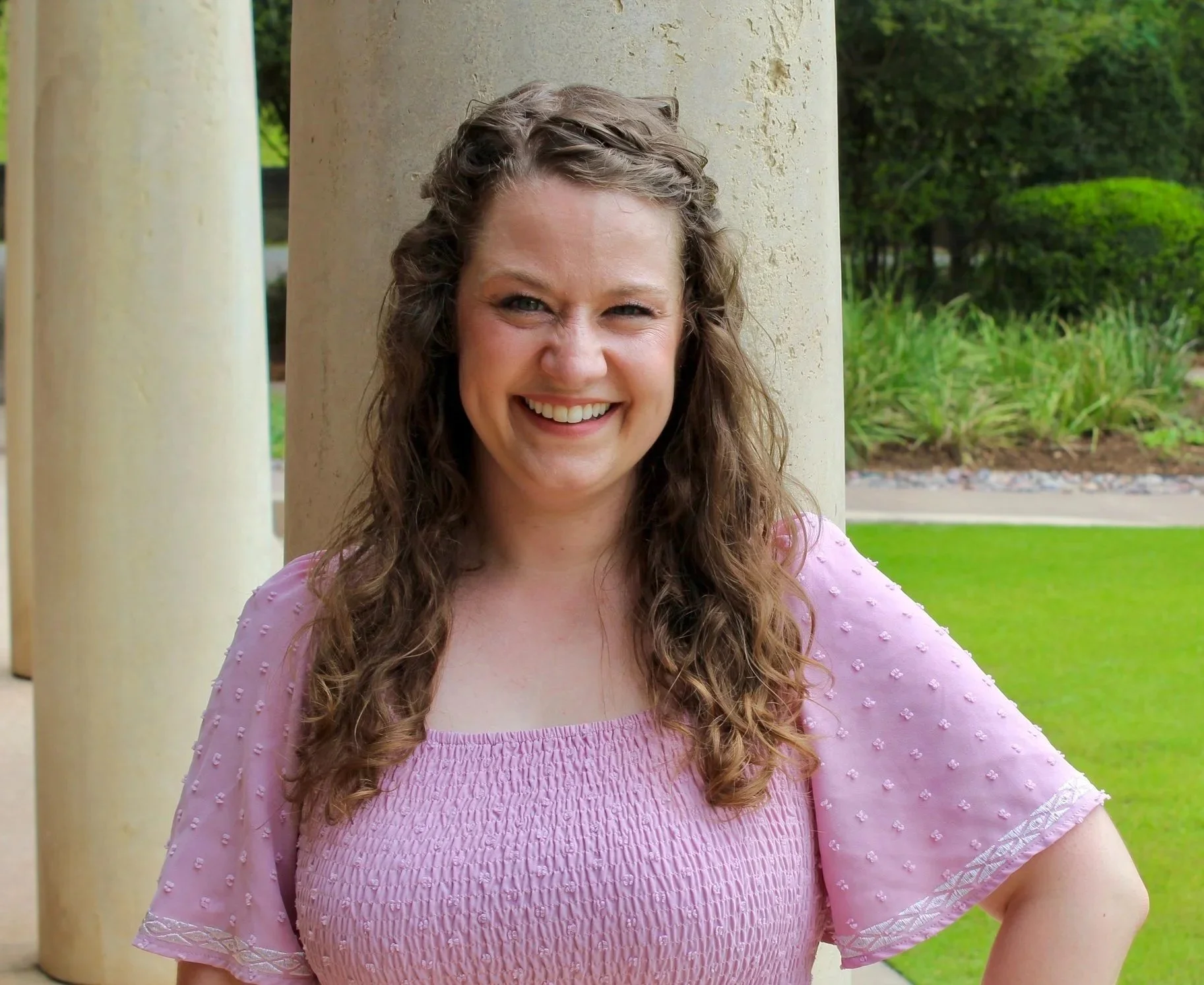 A woman with long curly hair smiling outdoors with a garden background, wearing a pink blouse with textured fabric and lace detail on the sleeves.
