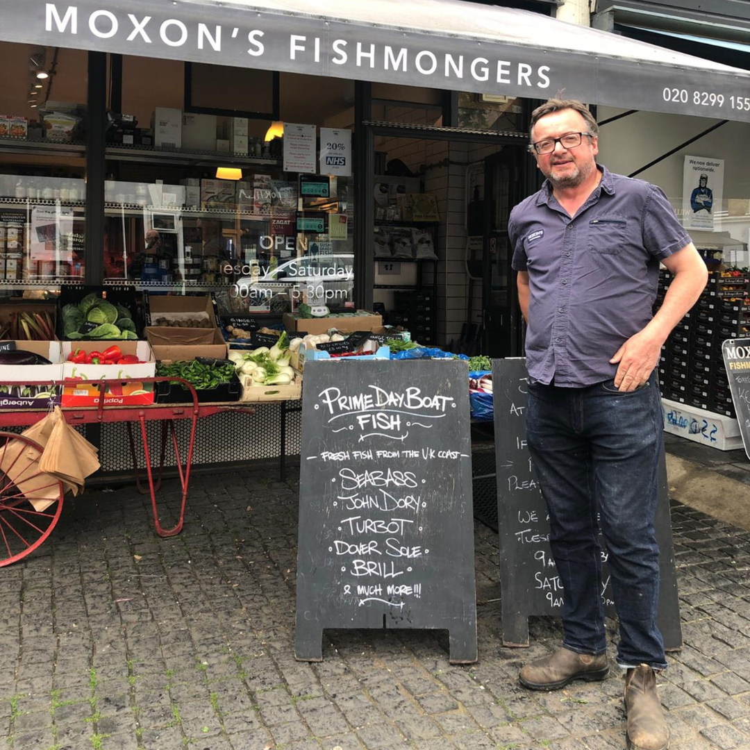 A man standing outside a fishmongers shop with a blackboard sign listing fresh fish and seafood. The shop has produce displayed outside and a glass window showing the interior. The shop name is Moxon's Fishmongers.