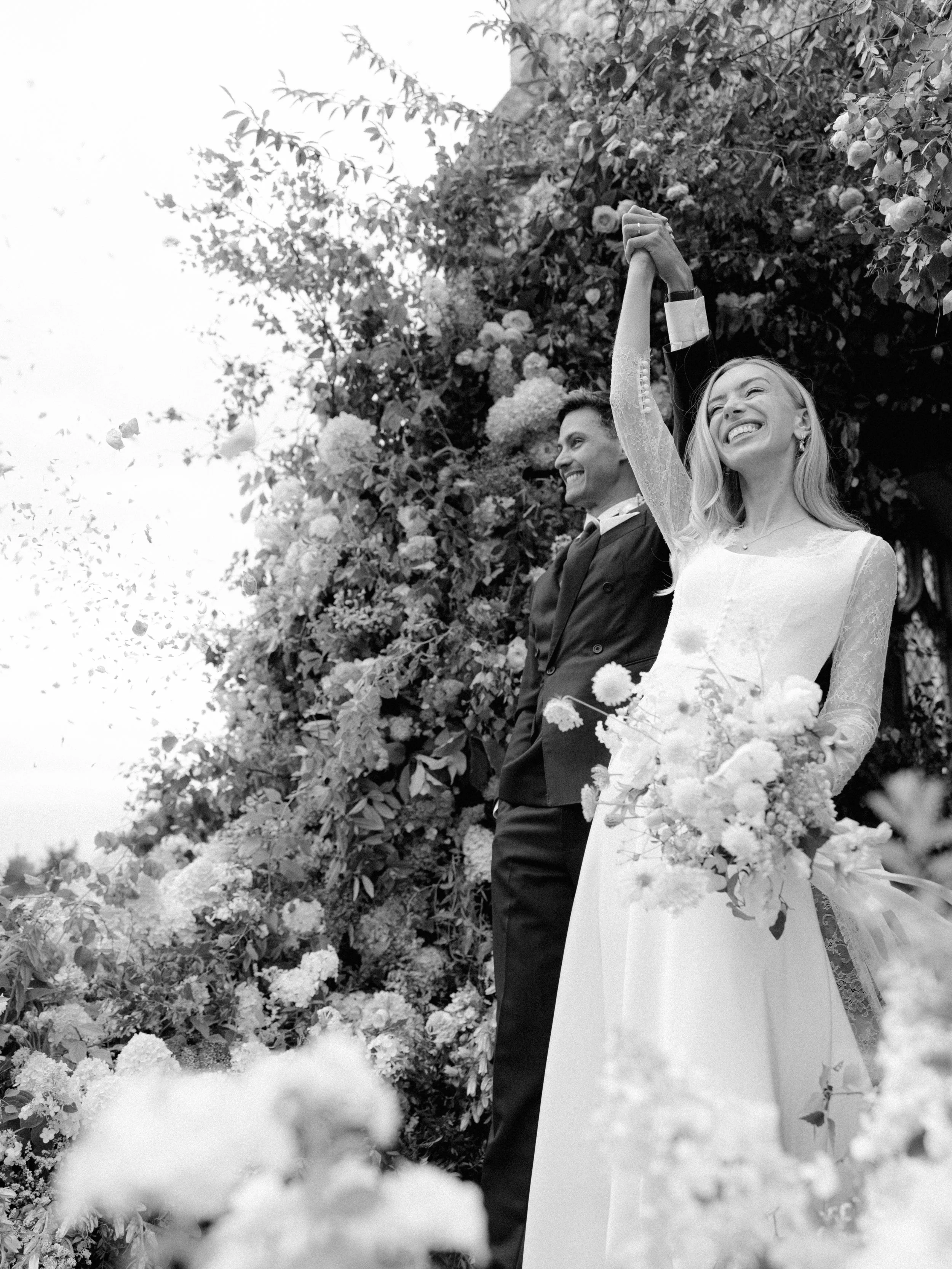A black-and-white photo of a bride and groom, smiling and holding hands raised in celebration, standing outdoors amidst flowers and greenery.