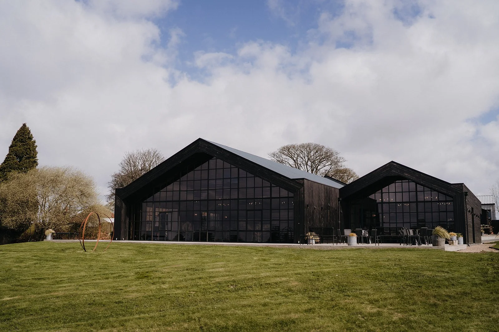 Modern black barn-style building with large glass windows, surrounded by a well-maintained green lawn and trees, under a partly cloudy sky.