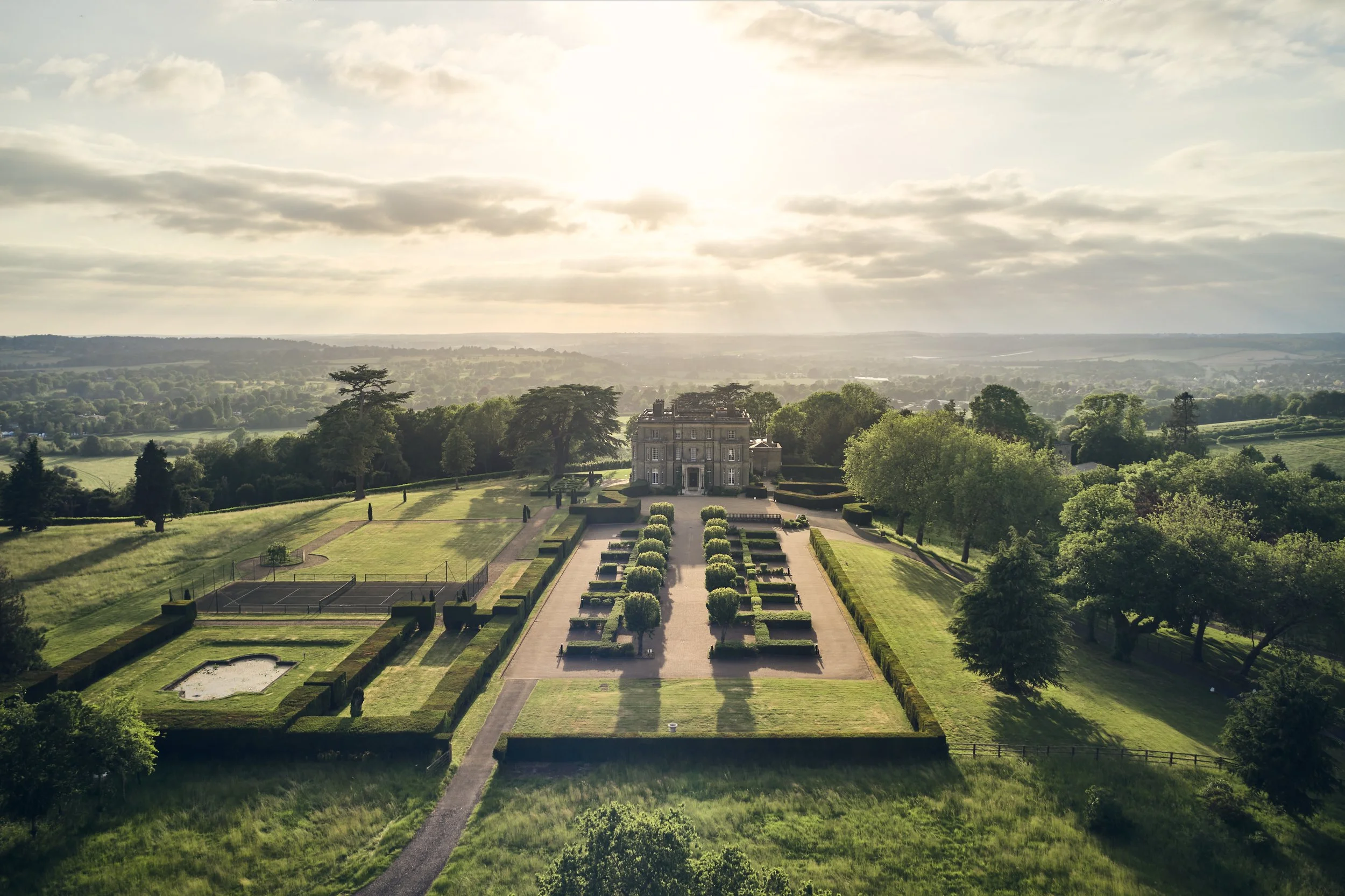 Aerial view of a large historic estate with a grand building at the center, surrounded by manicured gardens, trees, and lawns, under a partly cloudy sky with sunlight streaming through.