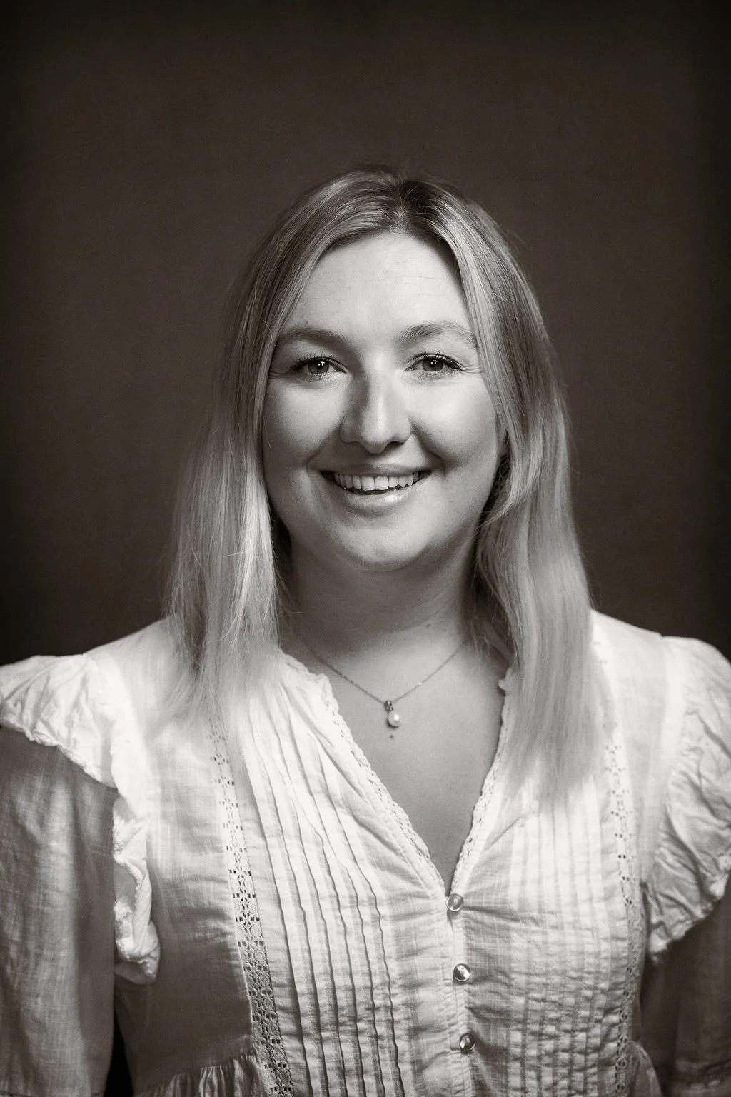 Black and white portrait of a smiling woman with shoulder-length hair, wearing a light-colored blouse with buttons and a necklace with a pendant.