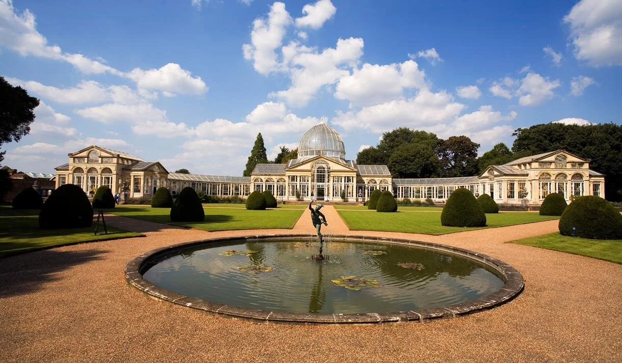 A glass conservatory with a domed roof, surrounded by well-manicured lawns, bushes, and a central fountain with a fountain sculpture, on a sunny day with a partly cloudy sky.