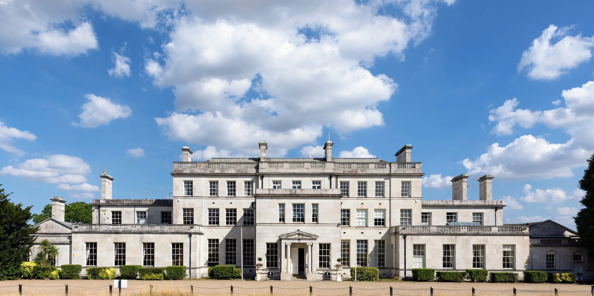 A large, elegant white stone mansion with a symmetrical facade, multiple windows, and a central entrance with a small porch. The mansion is surrounded by manicured bushes and trees under a bright blue sky with scattered white clouds.