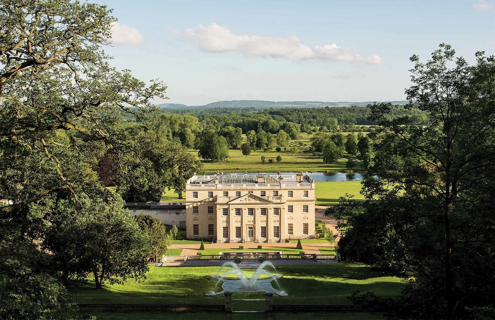 A grand historic mansion seen through trees, with a fountain in the foreground and lush green fields and distant hills in the background.