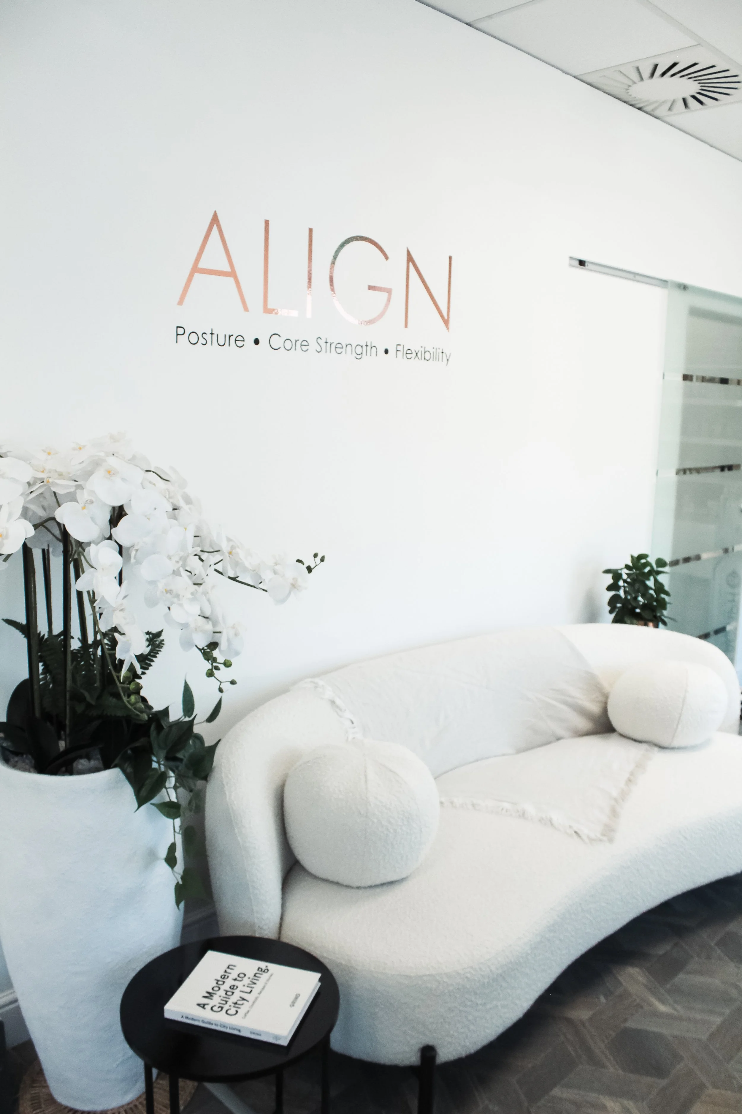 A modern, white, textured sofa with round cushions in a contemporary office or waiting area. A black side table with a book titled 'A Modern Guide to City Living' and a small black side table are nearby. There are white orchids in a large white pot o