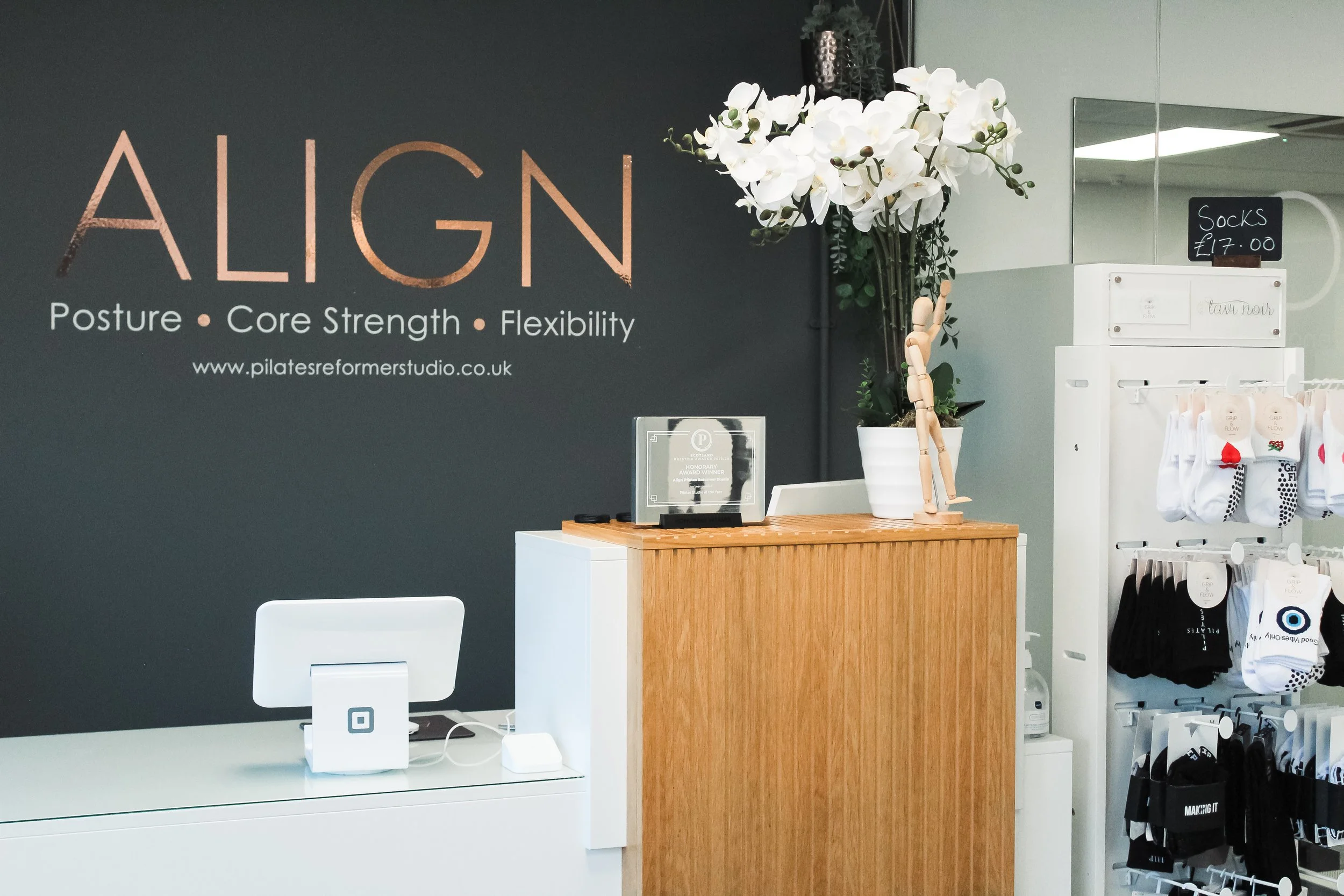 Interior of a yoga or Pilates studio reception area with a black wall displaying the studio's name 'ALIGN' and its focus on posture, core strength, and flexibility. There is a white orchid plant, a wooden reception desk with a small award or plaque, 