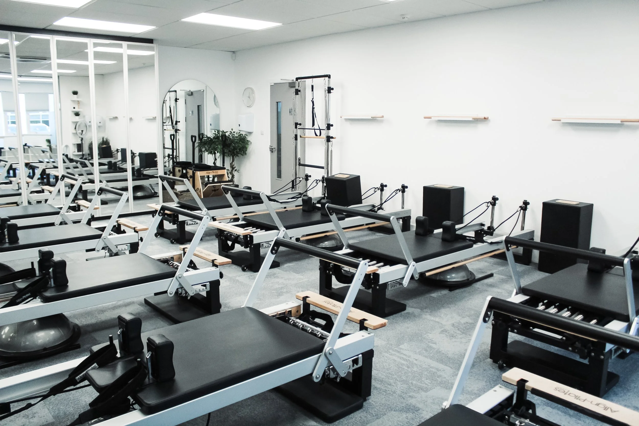 Fitness studio with reformer Pilates machines arranged in rows, a mirrored wall, and exercise equipment, white walls and ceiling lights.