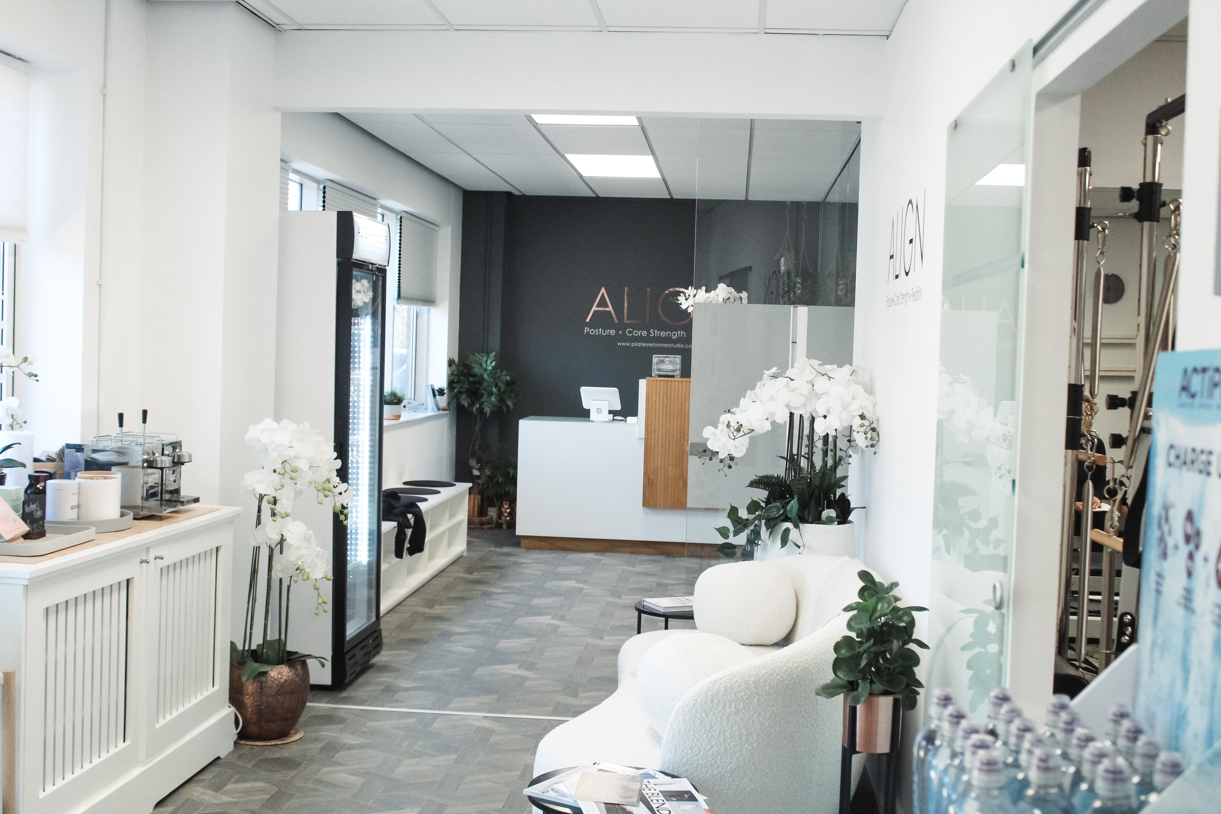 Interior of a modern office or wellness studio with white walls, plants, a reception desk, and a seating area. Visible signage on the wall reads 'ALIC' and 'Posture • Core Strength.'