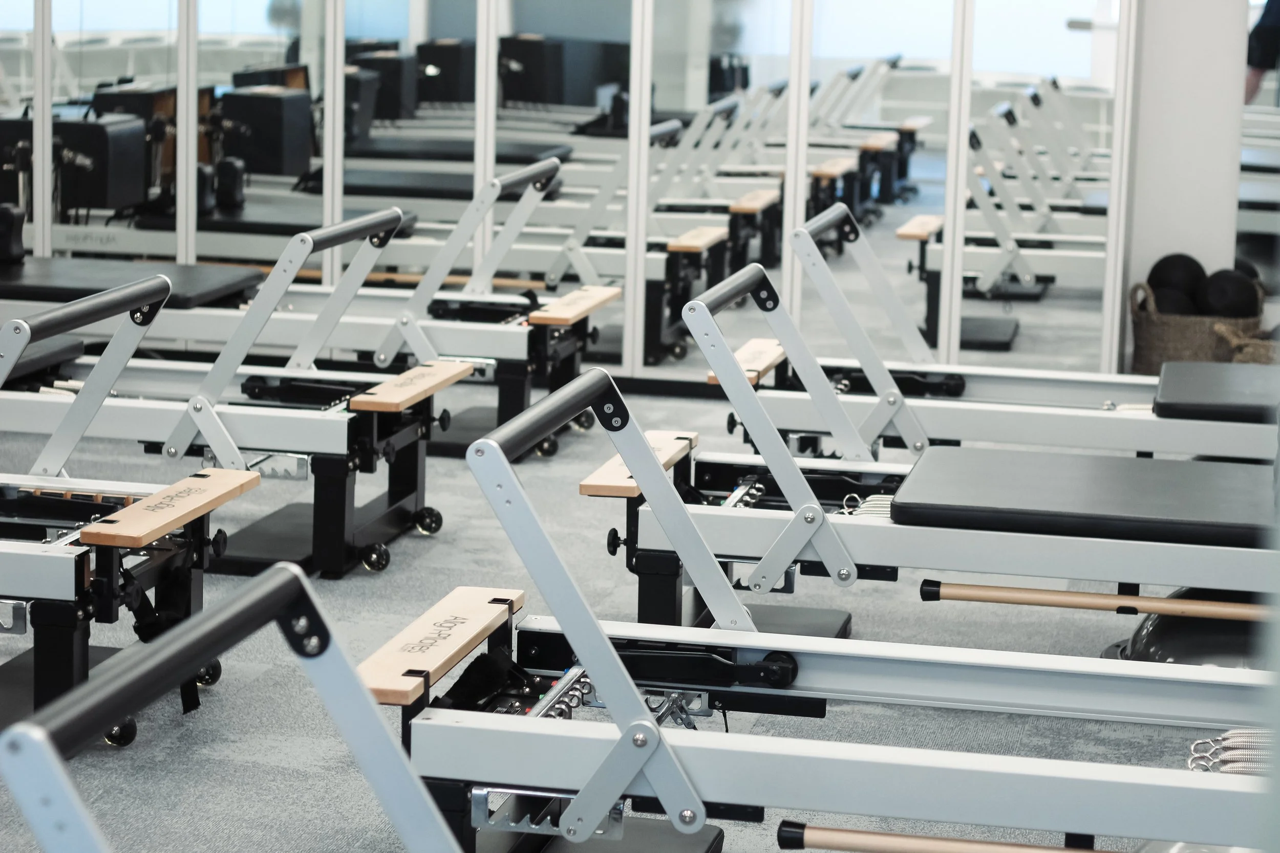 A fitness studio with multiple Pilates reformer machines arranged in rows, with a large mirror on the wall reflecting the equipment and part of a person in black clothing in the background.