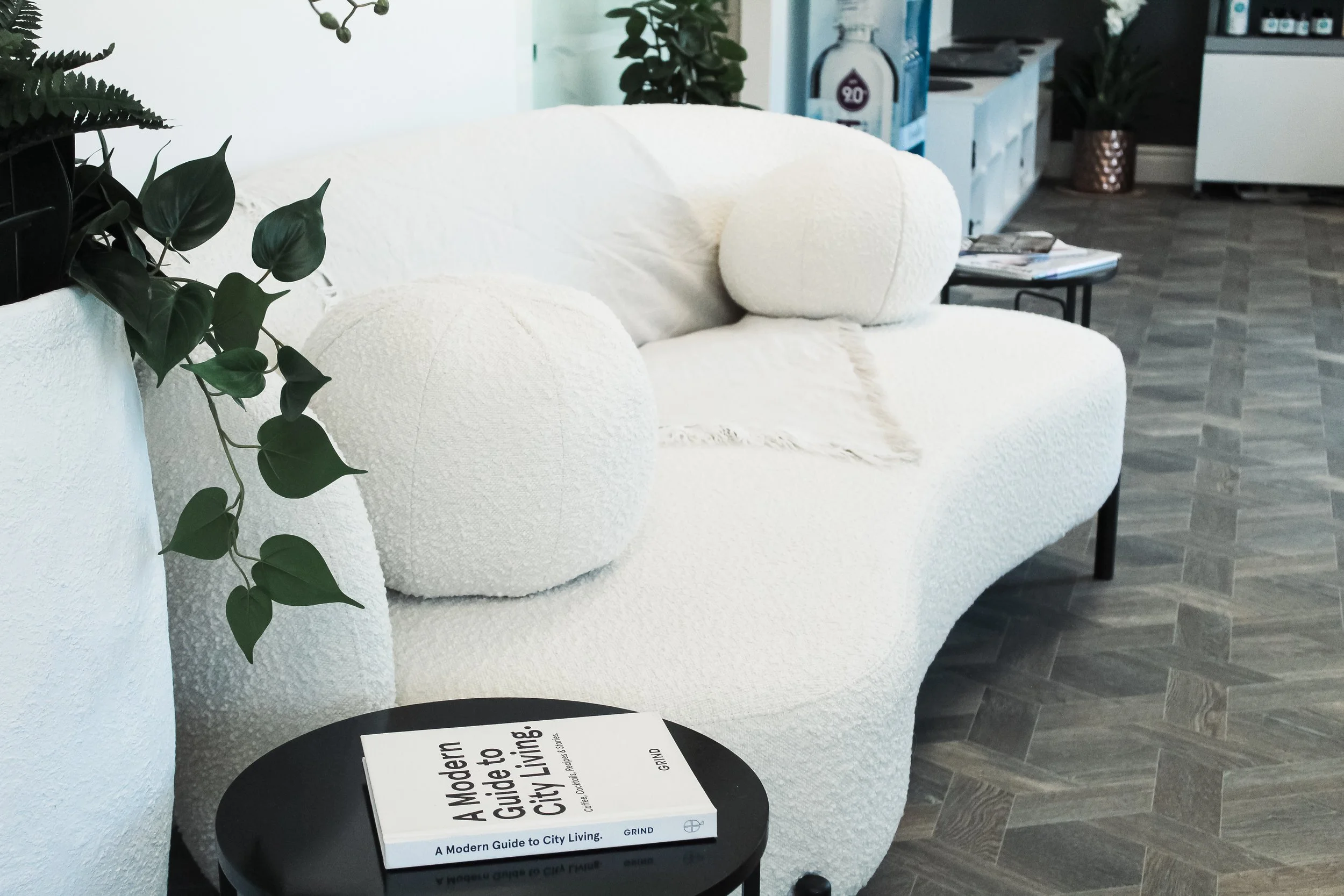 Close-up of a black table with a white book titled 'A Modern Guide to City Living' resting on it. Behind the table, there is a white textured sofa with round cushions, and a green leafy plant on the left side of the image. In the background, a modern