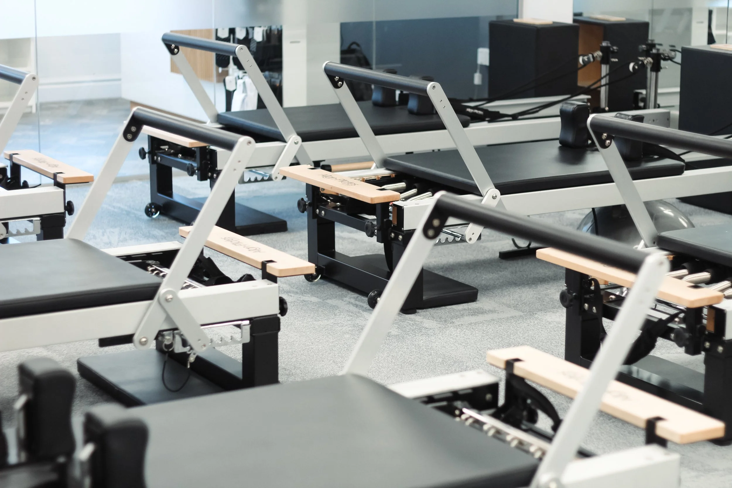Fitness studio with black Pilates reformer machines arranged in rows, mirrors on the wall, and a wooden floor.