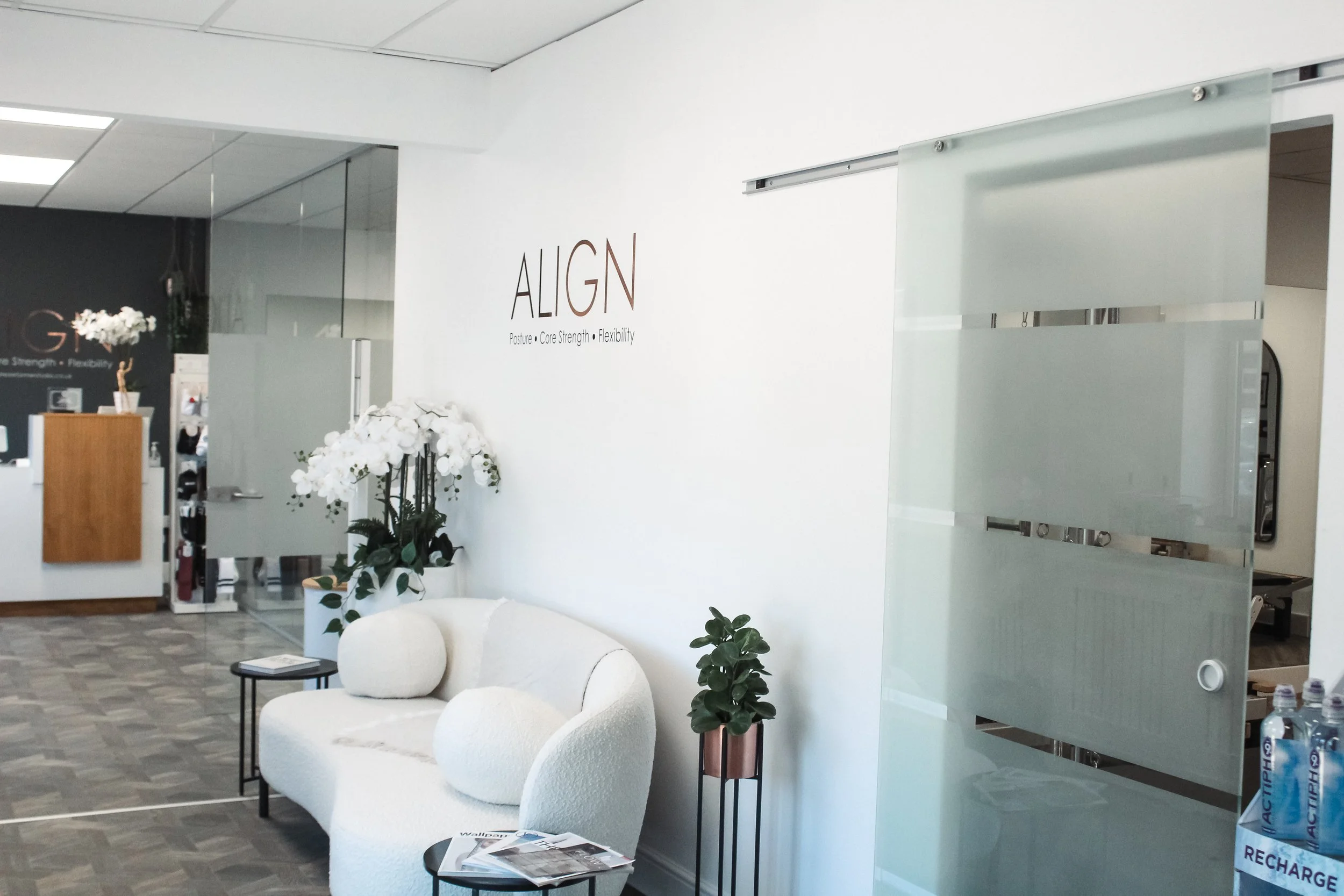 Interior of a modern office or reception area with white walls, glass partitions, a white curved sofa, potted plants, and a sign that reads 'ALIGN' with words 'Posture,' 'Core Strength,' and 'Flexibility' underneath.