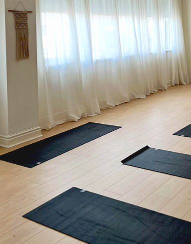Dark yoga mats arranged on a light wooden floor in front of large windows with white curtains in a bright room.
