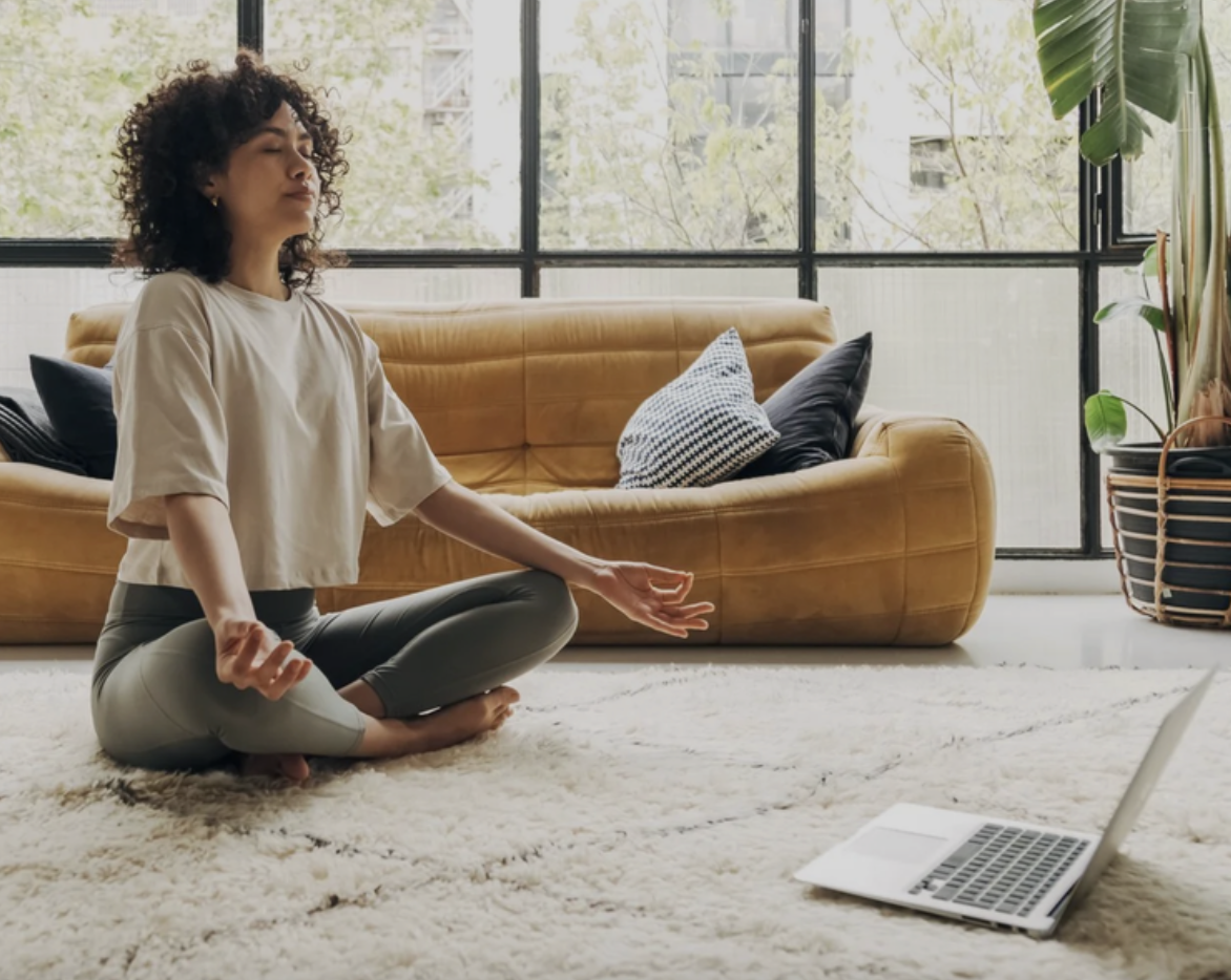 A laptop on a small round table displaying a video of a person practicing yoga in a room with angled ceilings and large windows.