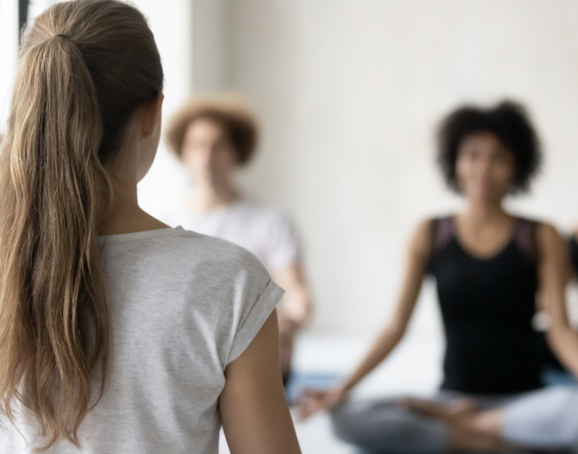 A woman with long brown hair in a ponytail, wearing a light gray T-shirt, seen from behind, participating in a group meditation or yoga session with two women in the background, one with curly brown hair and the other with curly black hair, sitting on mats in a well-lit room.