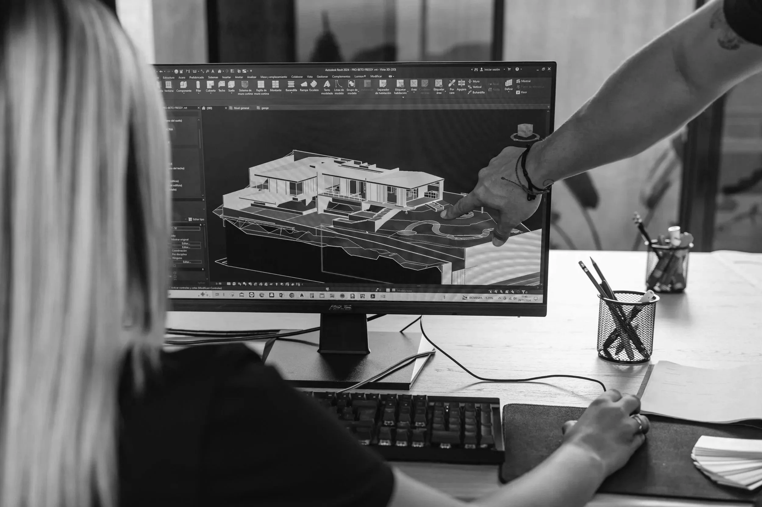 A person points at a 3D architectural house model displayed on a computer screen, with another person sitting nearby. The scene is in a workspace with office supplies on the desk.