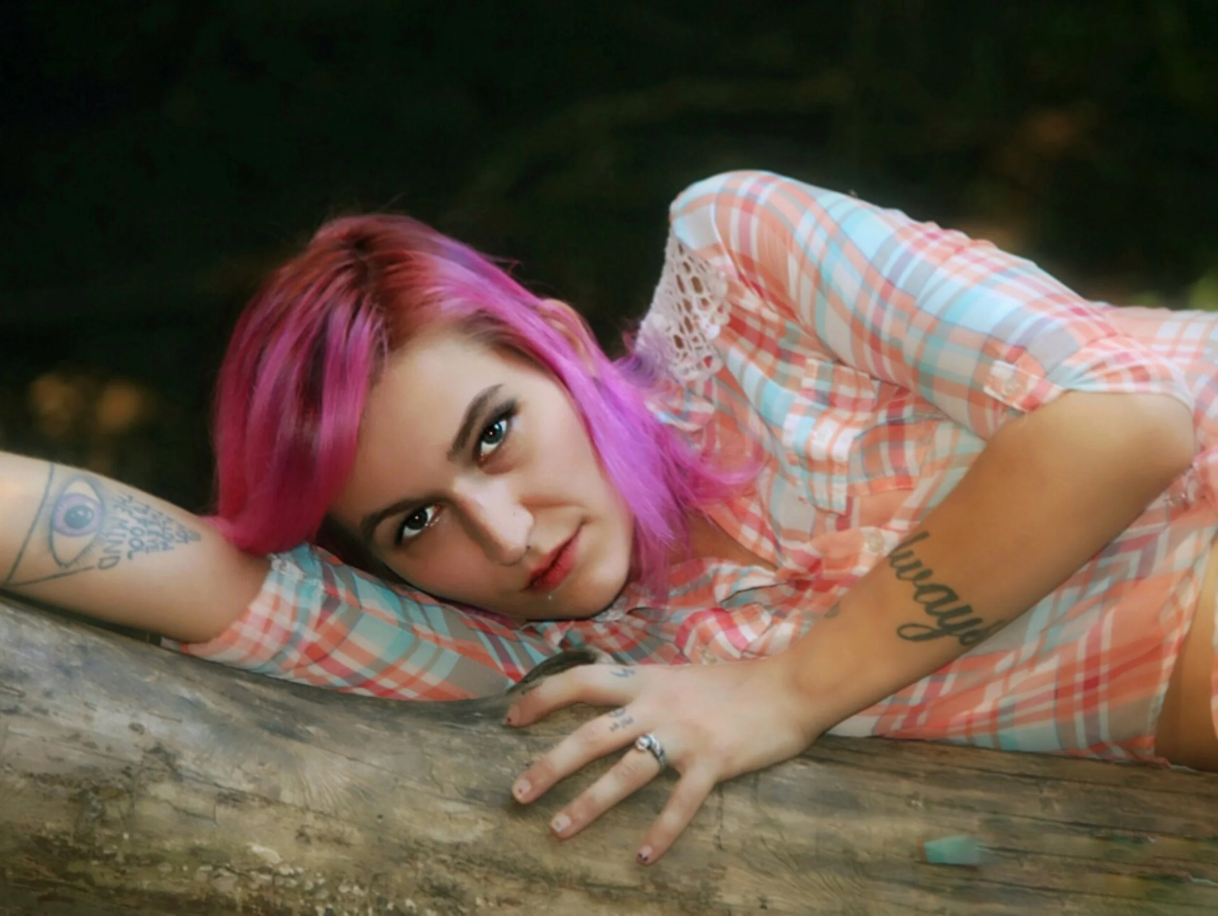 A striking portrait of a woman with vibrant pink hair, lying on a weathered tree trunk. She looks directly at the camera with a relaxed expression, wearing a light plaid shirt and showcasing unique arm tattoos. The soft, blurred background enhances t