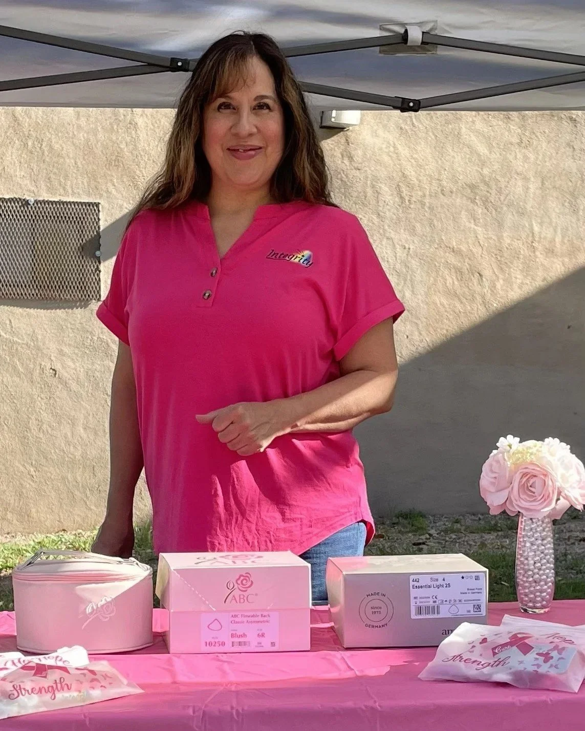 A woman wearing a pink shirt standing behind a table with pink boxes, a pink vase with flowers, and pink items, outdoors under a canopy.