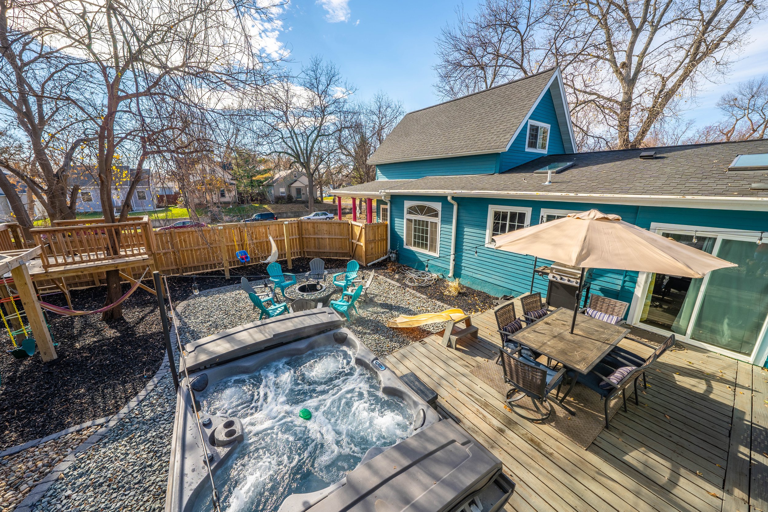 A backyard view featuring a jacuzzi, wooden deck with outdoor dining set and umbrella, outdoor seating area with fire pit, hammock, playground with slide, and a blue house. Trees and a wooden fence are visible in the background.
