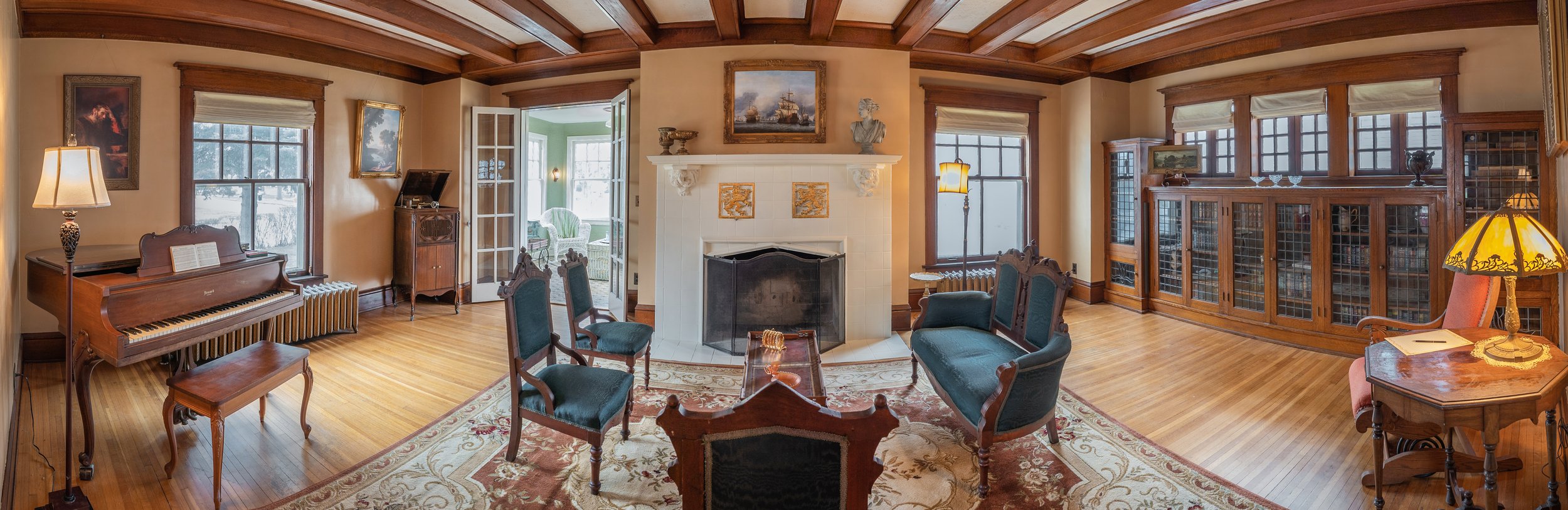 Vintage living room with a grand piano, antique furniture, and a fireplace. The room features a hardwood floor, a decorative rug, and a coffered ceiling. There are large wooden cabinets with glass doors, a lamp, and framed artwork on the walls. A sun
