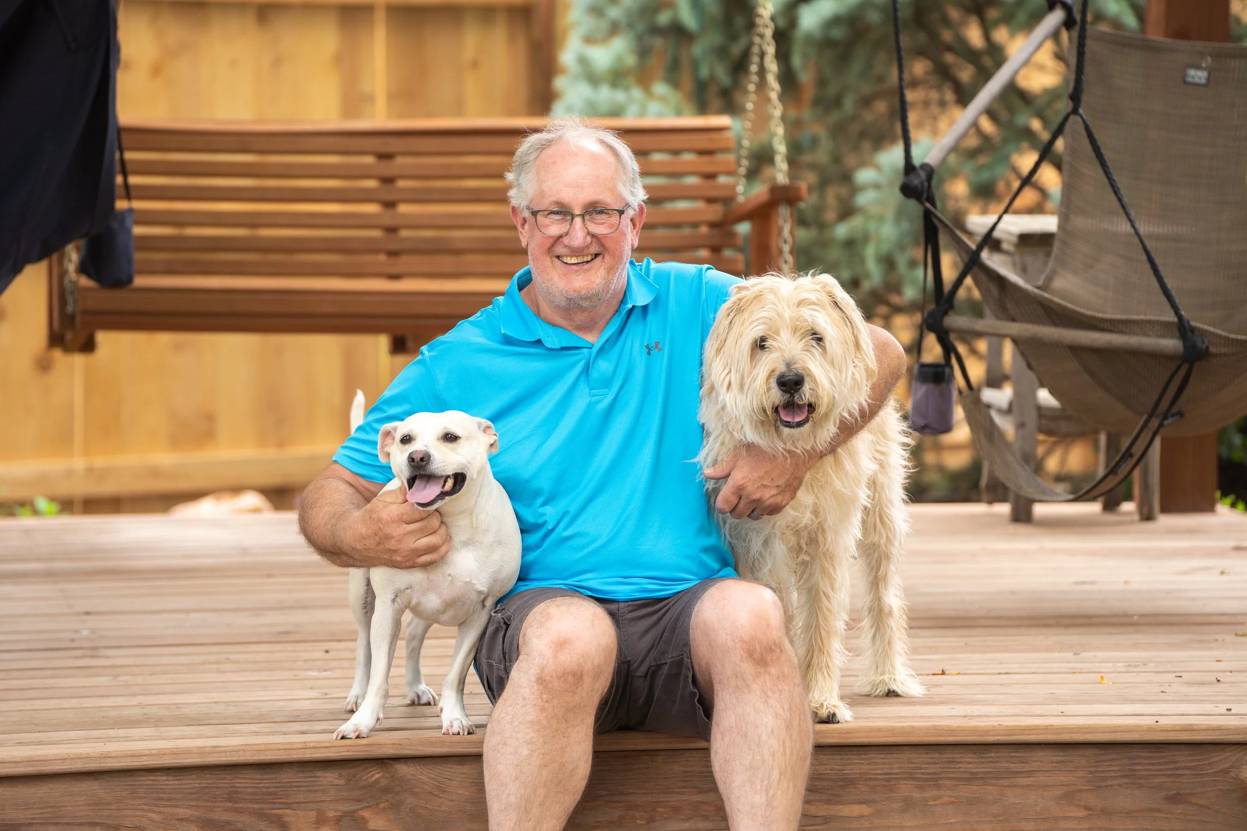 A man sitting on a porch with two dogs, a small white one and a larger fluffy one, smiling for the photo.