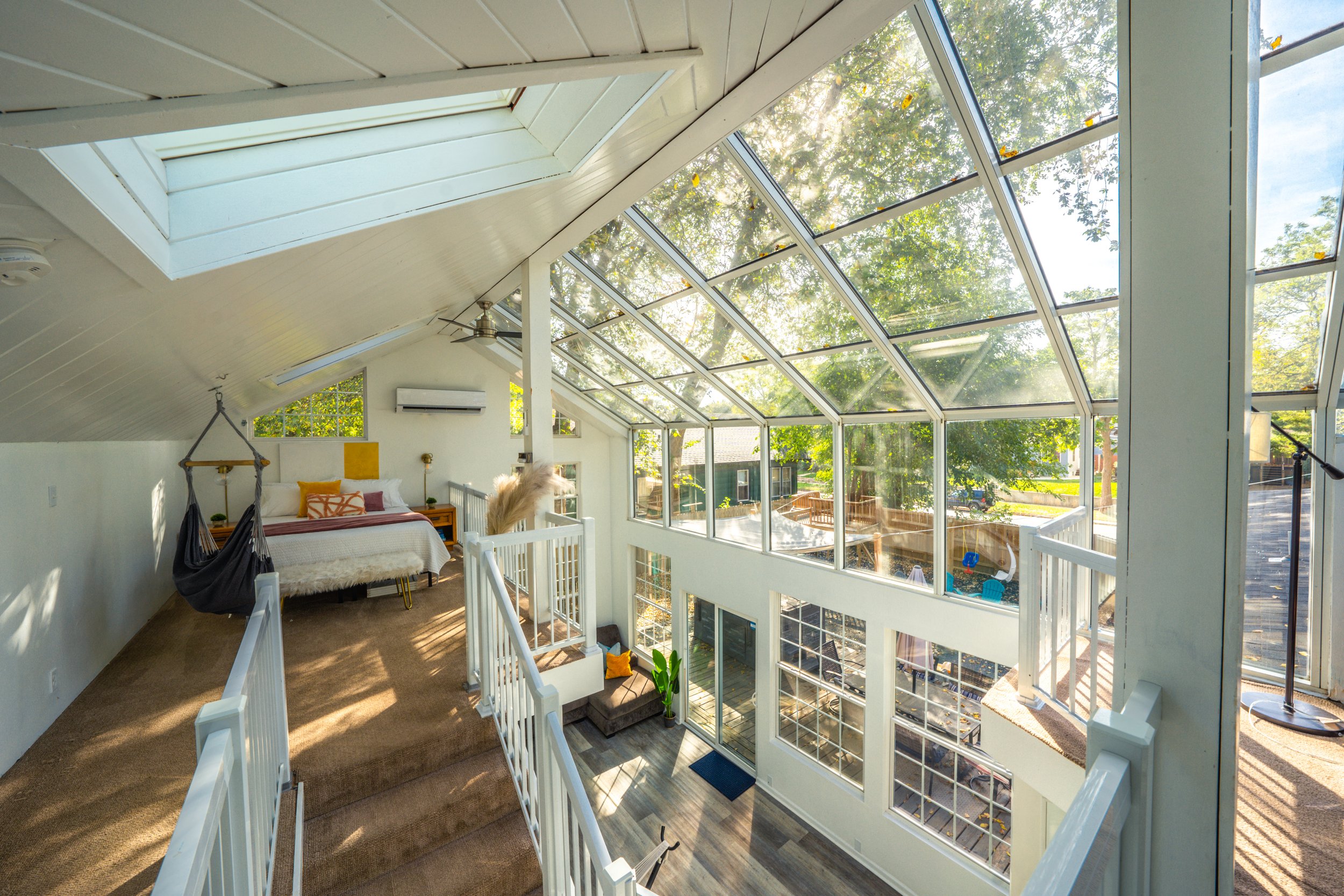 Modern interior with a loft bedroom, skylight windows, and glass walls overlooking a patio and trees; features include a hammock chair, cozy bed with colorful pillows, and a split air conditioner.