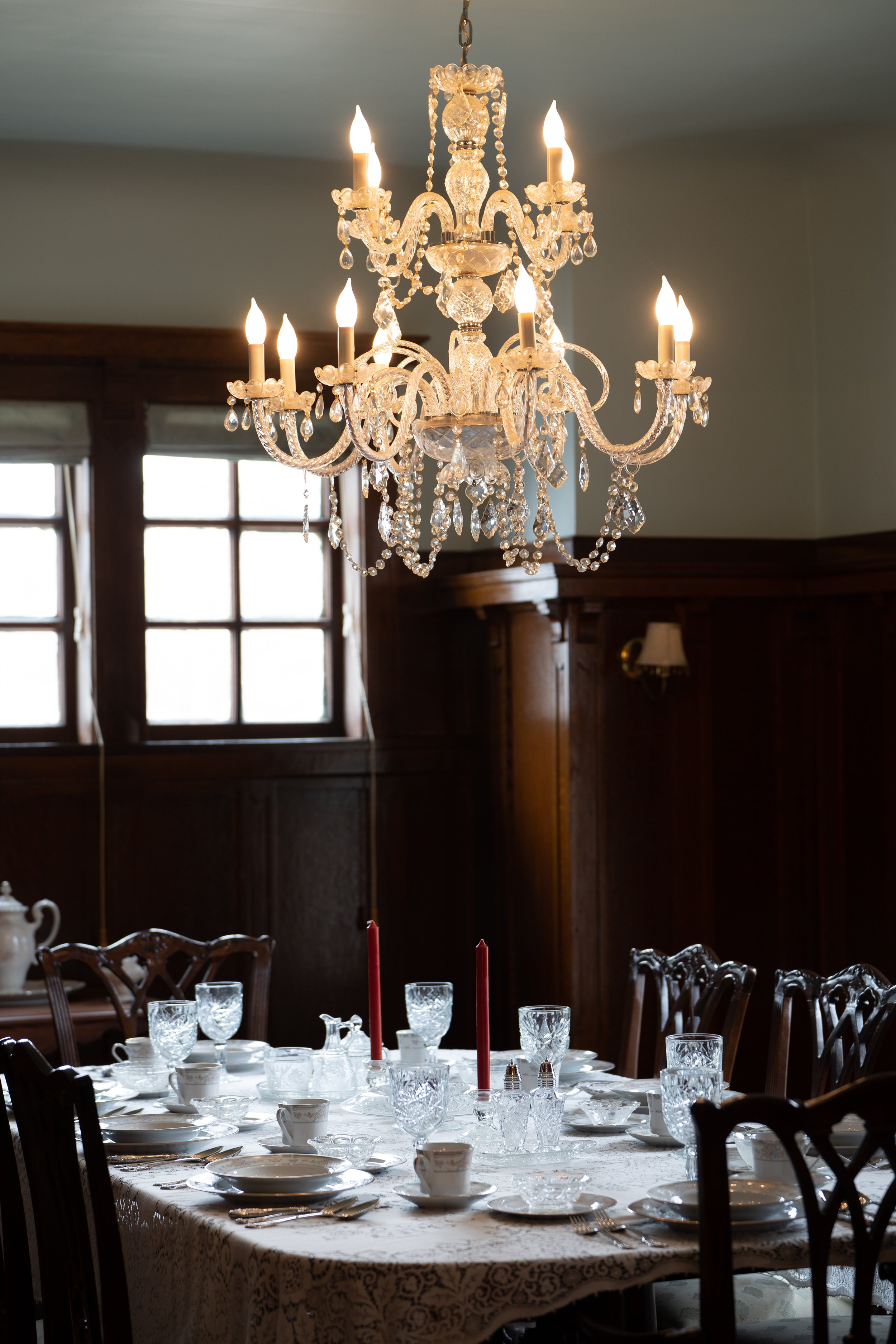 Elegant dining room with crystal chandelier, set dining table with white tableware and red candles, wooden chairs, and wooden paneling.