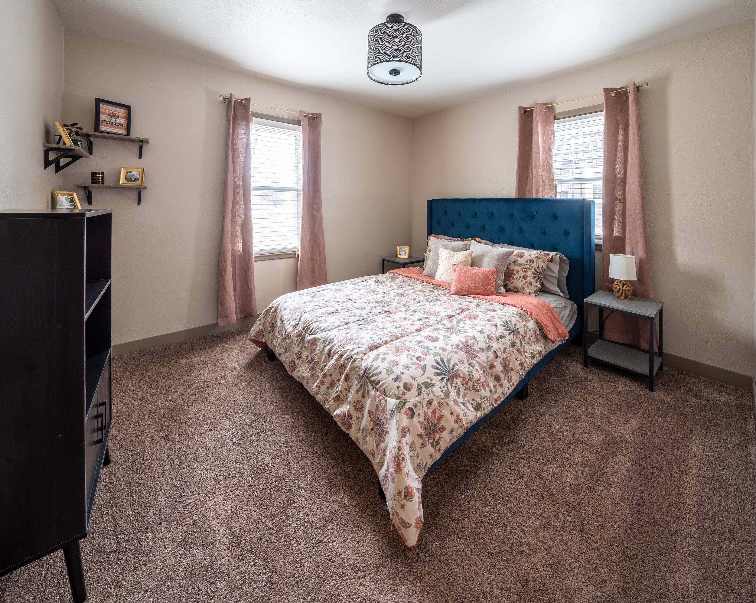 A cozy bedroom with a floral-patterned bedspread on a bed with a blue tufted headboard. The room features two windows with pink curtains, a carpeted floor, and side tables with lamps. Shelves with small decorations are mounted on the wall.
