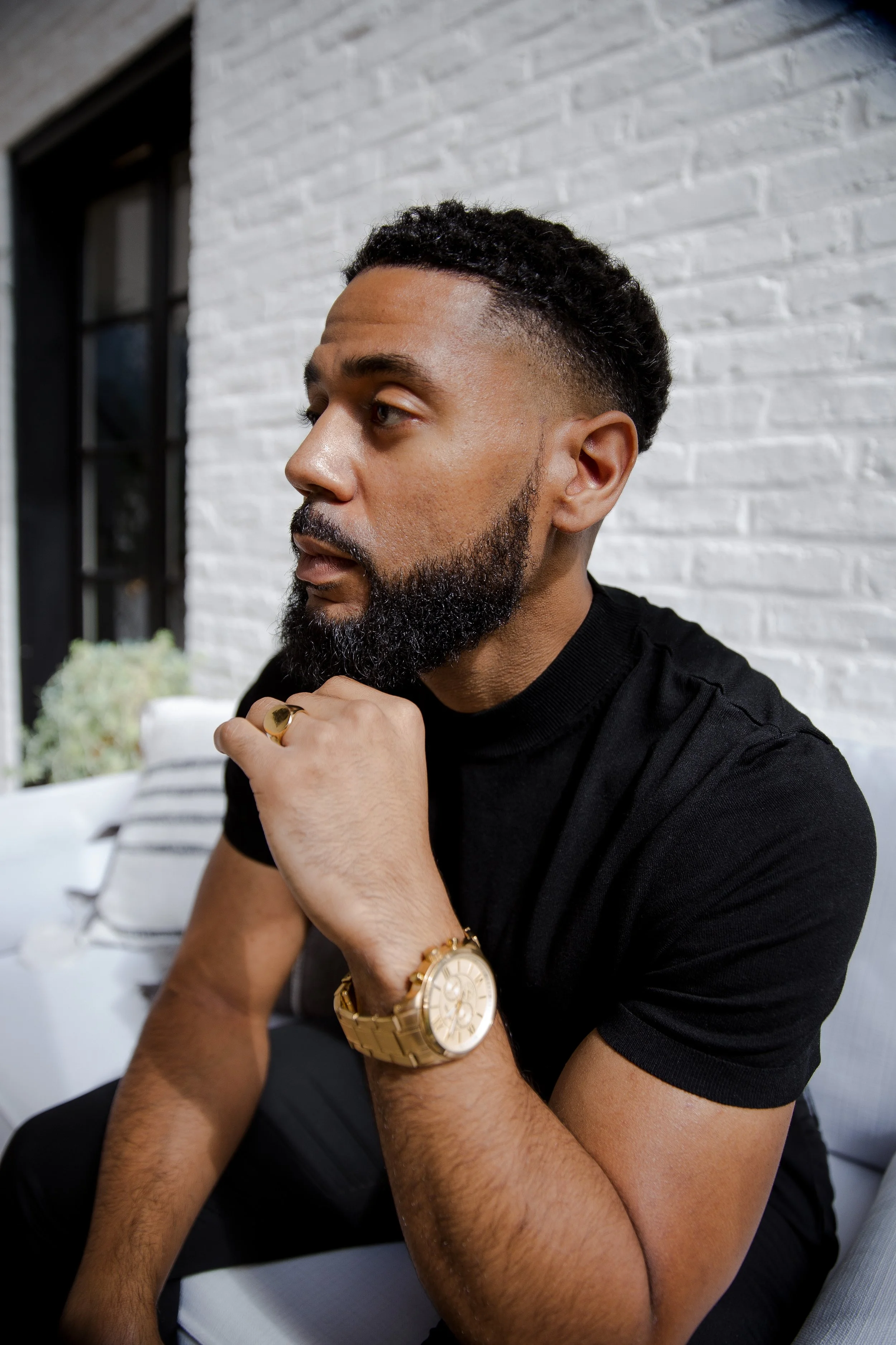 A man with a beard and mustache wearing a black t-shirt, gold watch, and gold ring, sitting with his hand on his chin, looking to the side against a white brick wall.