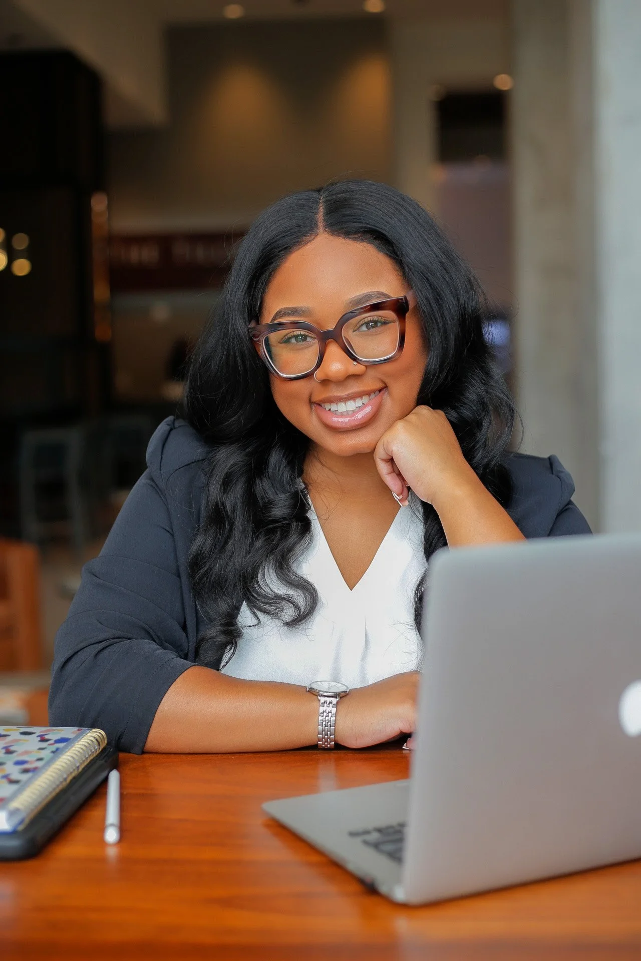 A smiling woman with glasses sitting at a wooden table in a cafe, using a silver MacBook, with a notebook and pen beside her.