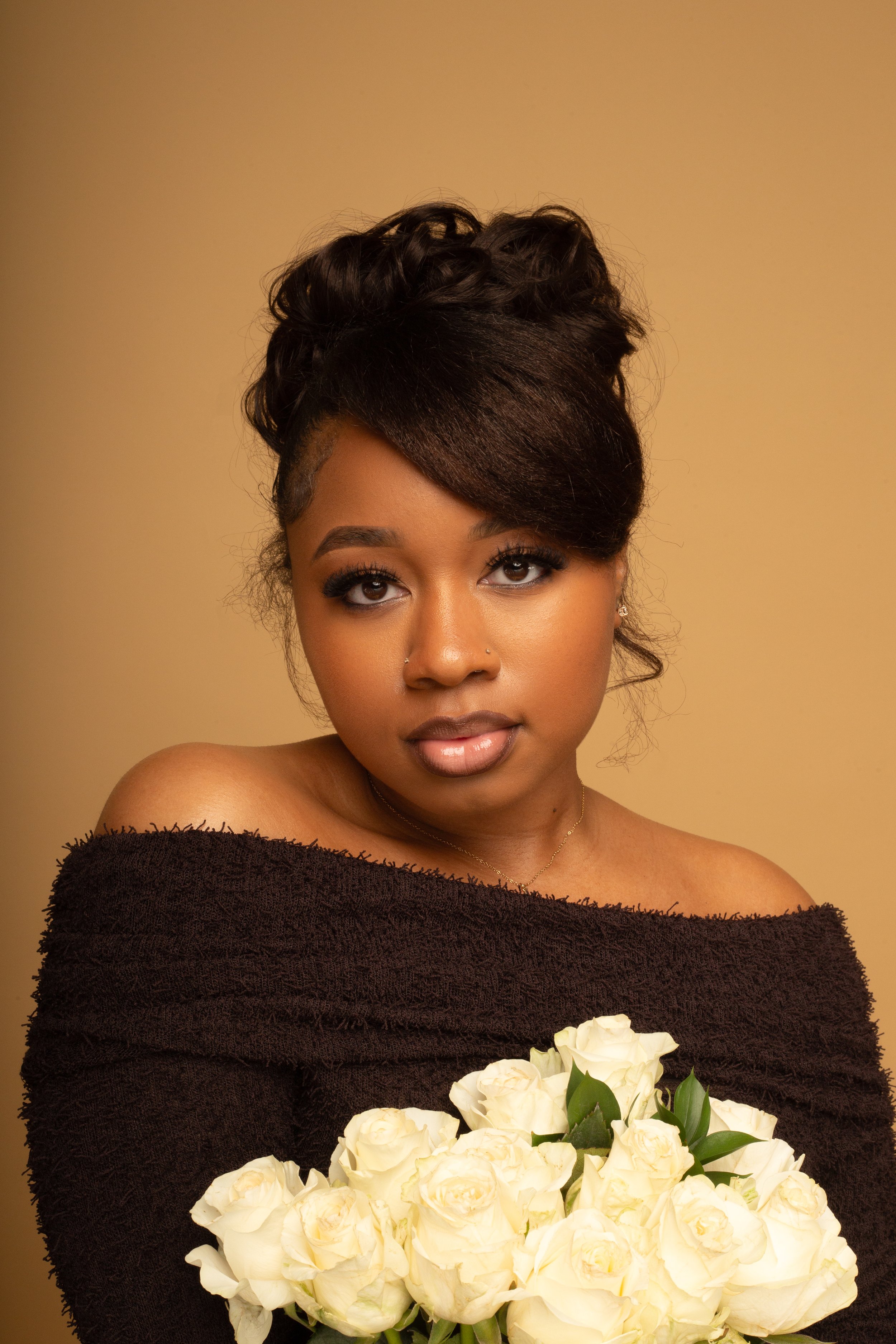 Portrait of a young woman with styled hair holding white roses, wearing a black off-the-shoulder top against a beige background.