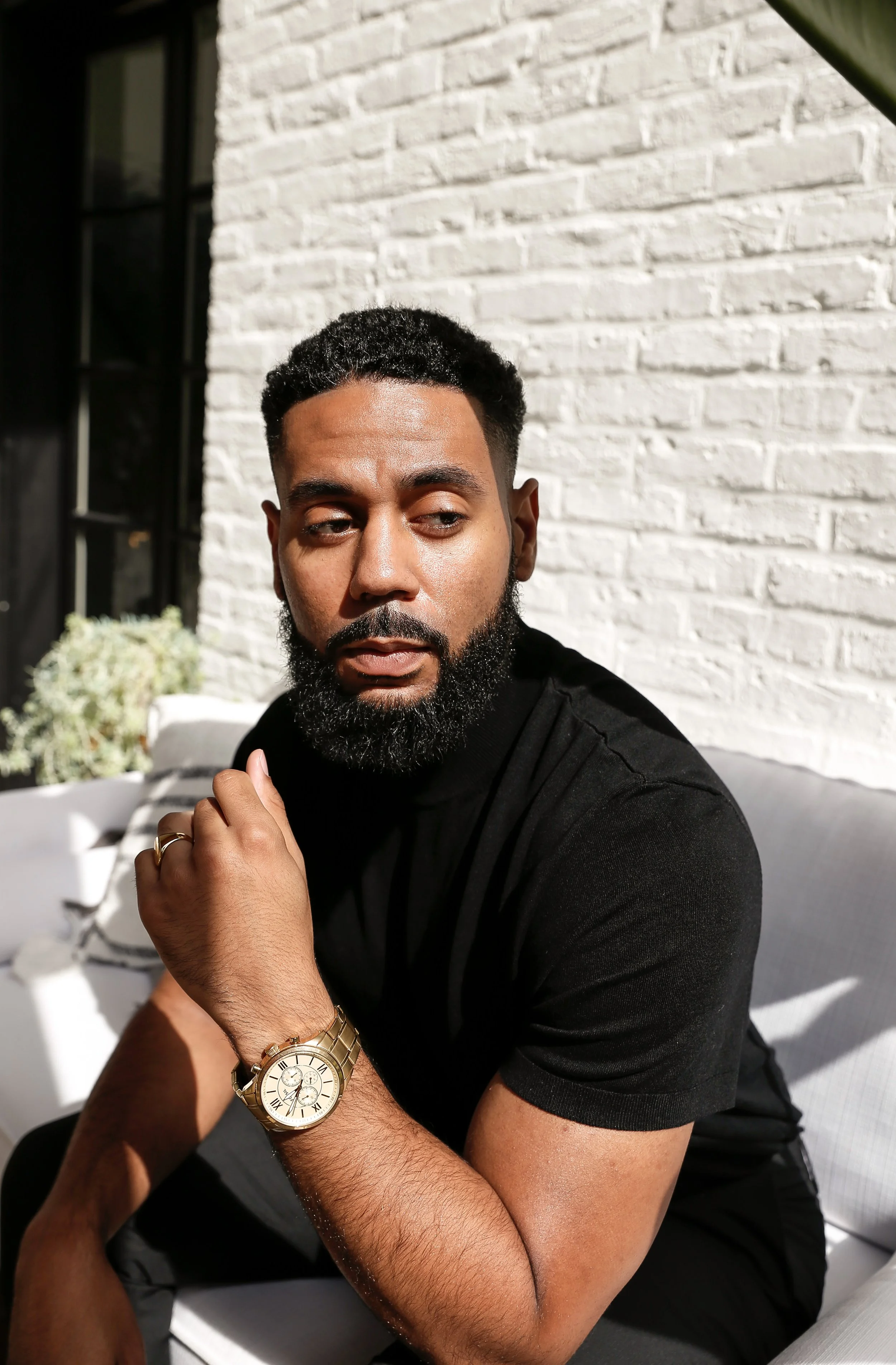 A man with a beard sitting outside on a white couch, wearing a black shirt and a gold watch, with a white brick wall and sunlight behind him.