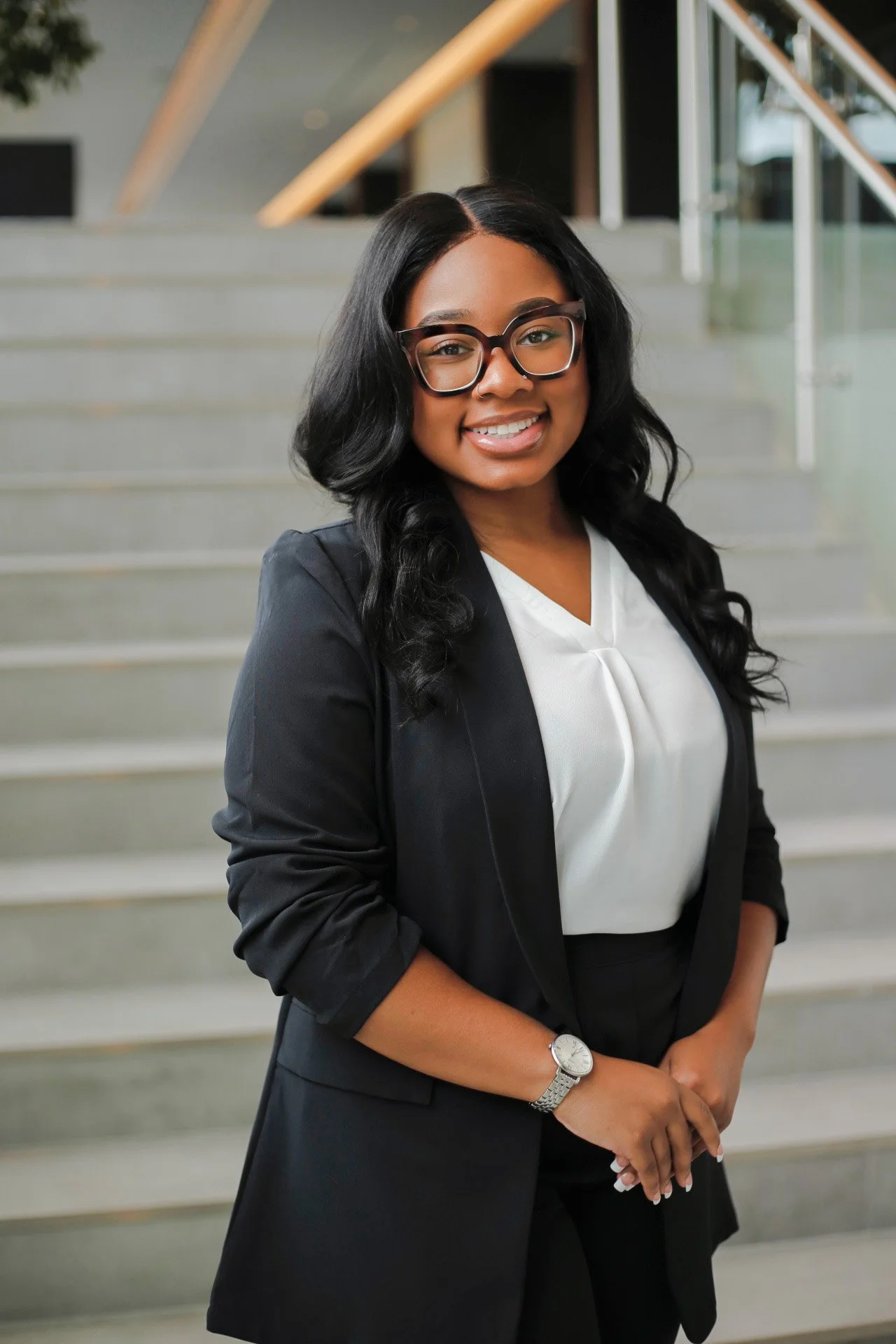 A woman with long black curly hair, glasses, and a bright smile, dressed in a black blazer and white blouse, standing on a staircase in a modern indoor setting.