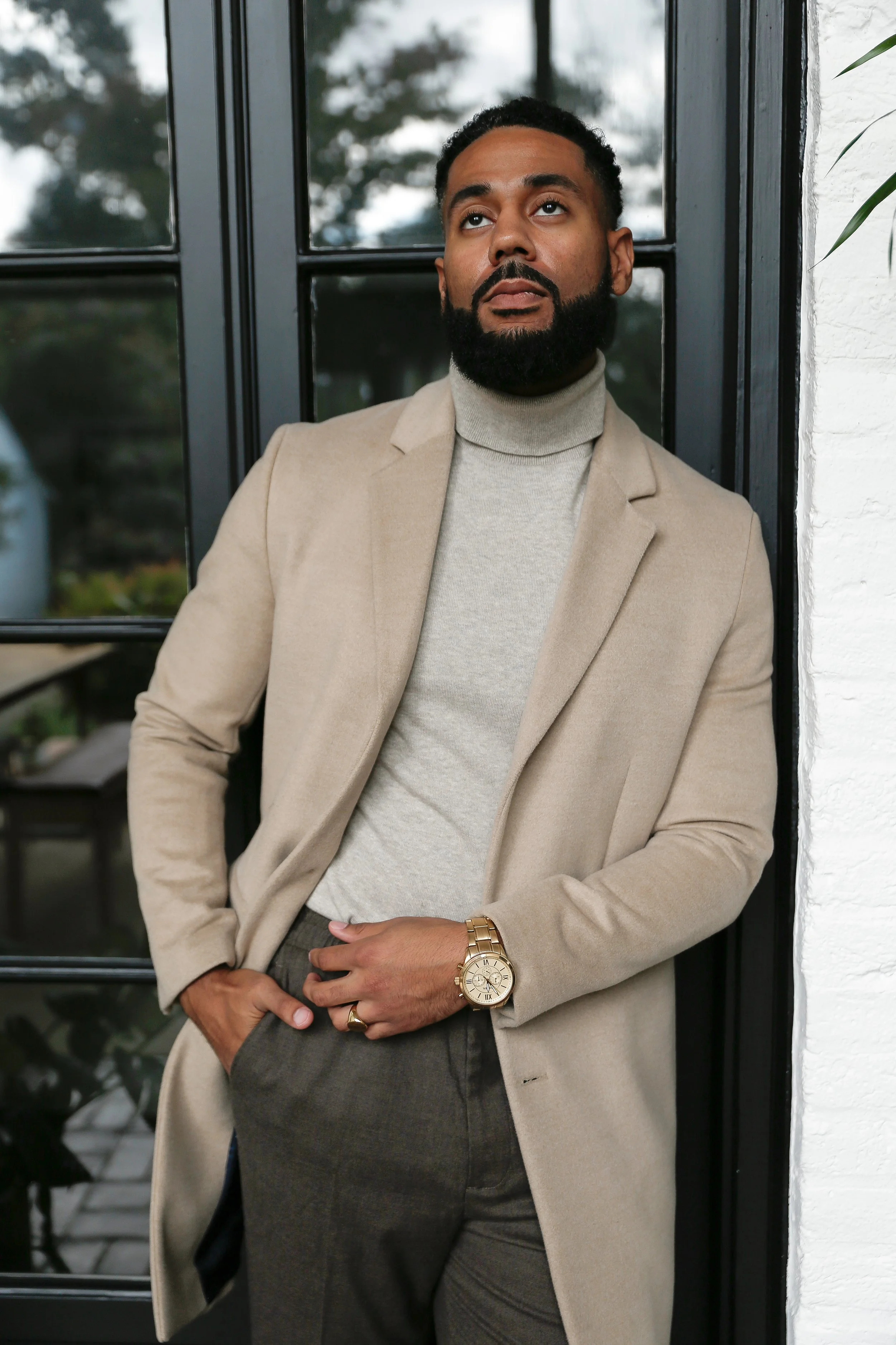 A man with a beard wearing a beige coat, light gray turtleneck, and dark pants, leaning against a black-framed window with greenery visible in the background.