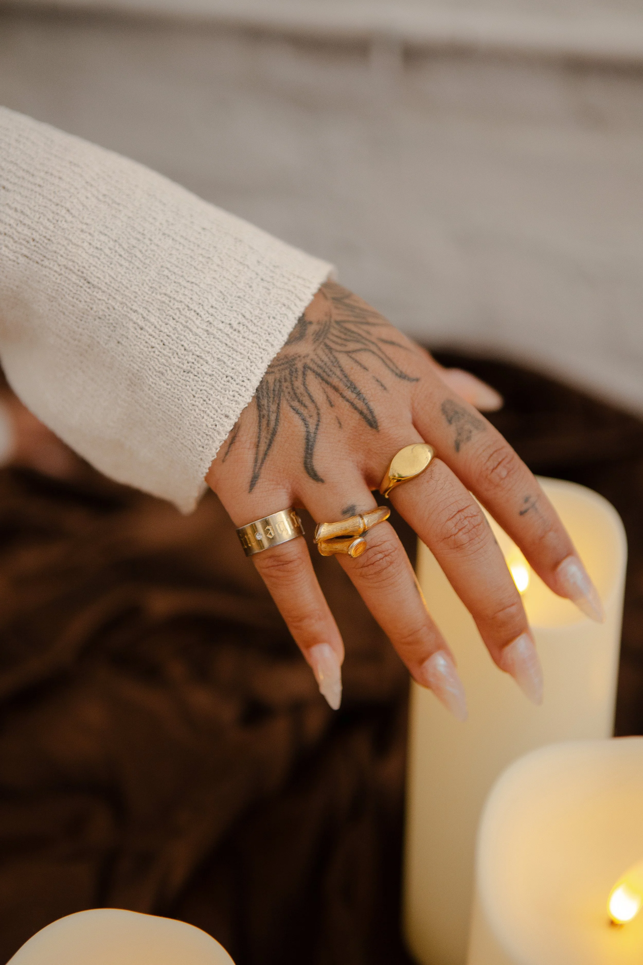 A person's hand resting on a white candle, adorned with multiple gold rings, with visible tattoos on fingers and back of the hand, and a beige sweater sleeve partially covering the wrist.