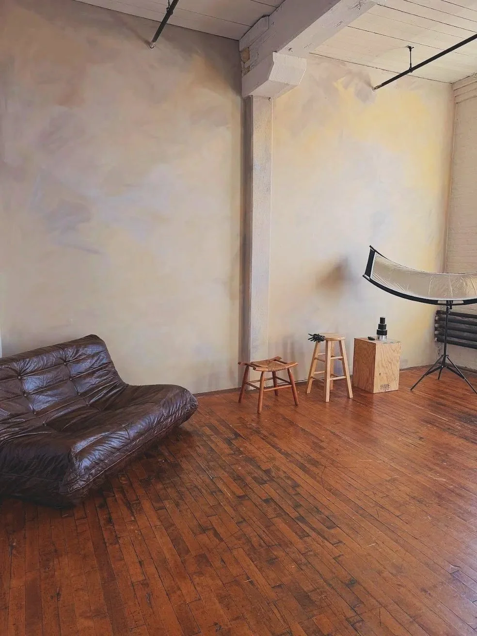 Empty room with wooden floor, beige wall, brown leather couch, two wooden stools, a wooden box, and photography equipment including a softbox and a backdrop stand.