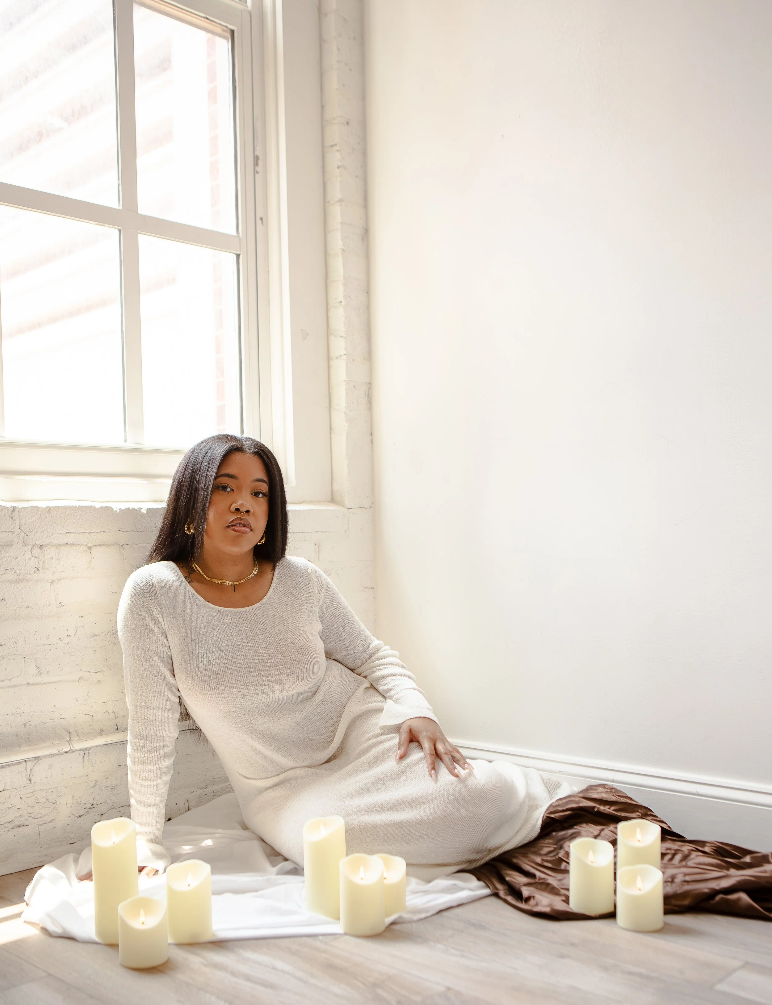 A woman with dark hair sitting on the floor near a window, surrounded by lit candles, wearing a white long-sleeve dress.
