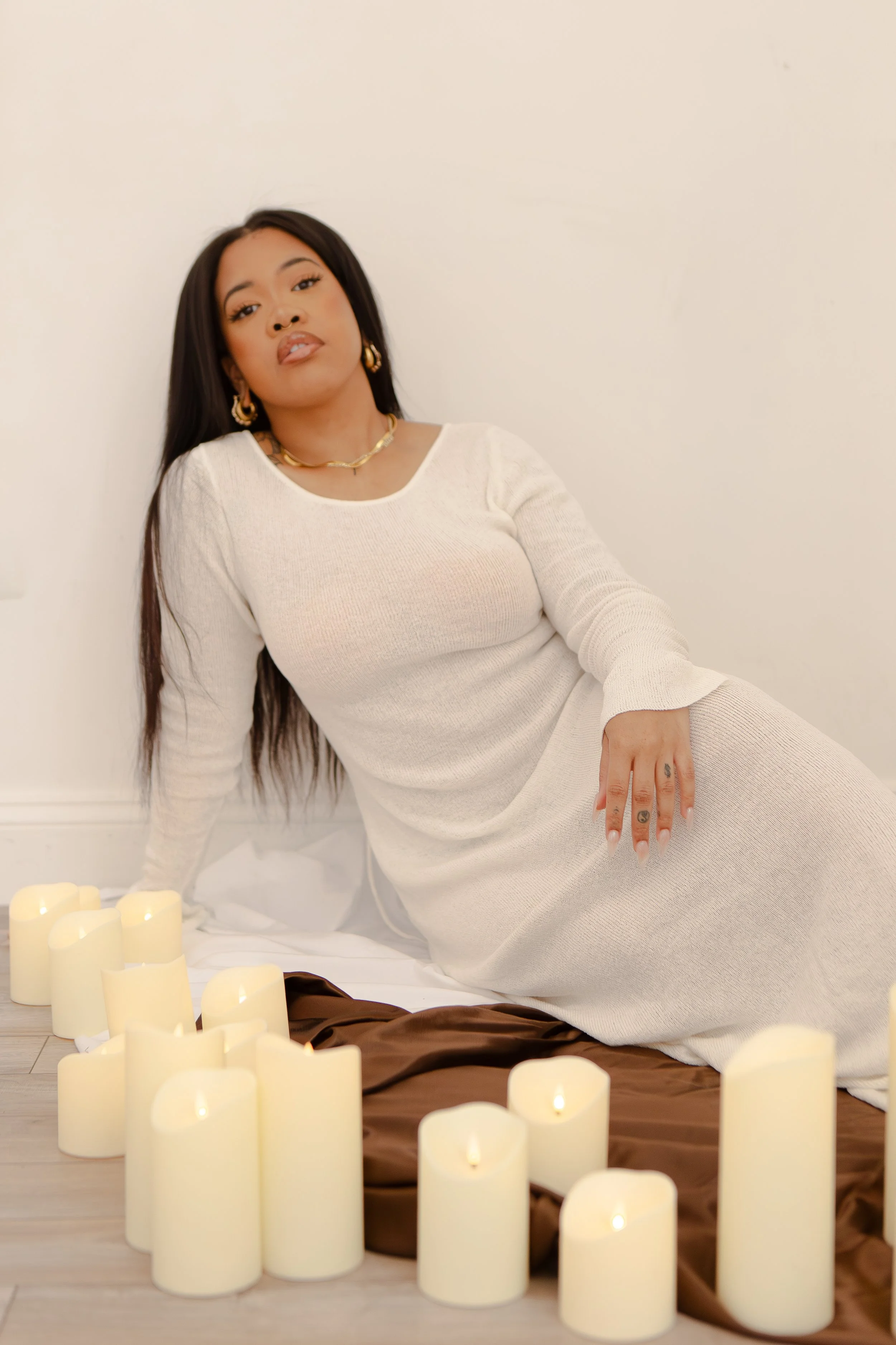 A woman with long dark hair, wearing a white dress and gold jewelry, sitting on the floor surrounded by lit white candles and a brown satin sheet.