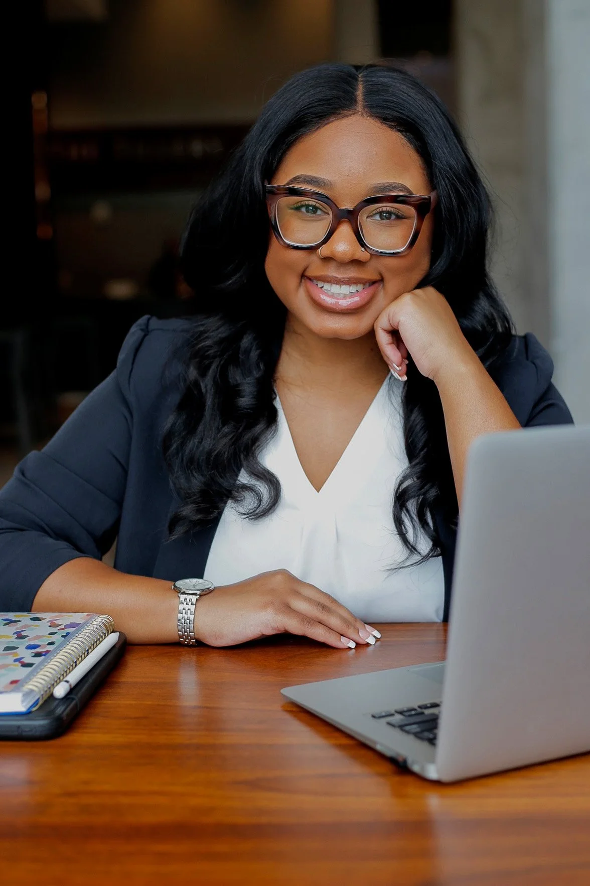 A woman with black hair, glasses, and a nose piercing smiling at a laptop in a professional setting.