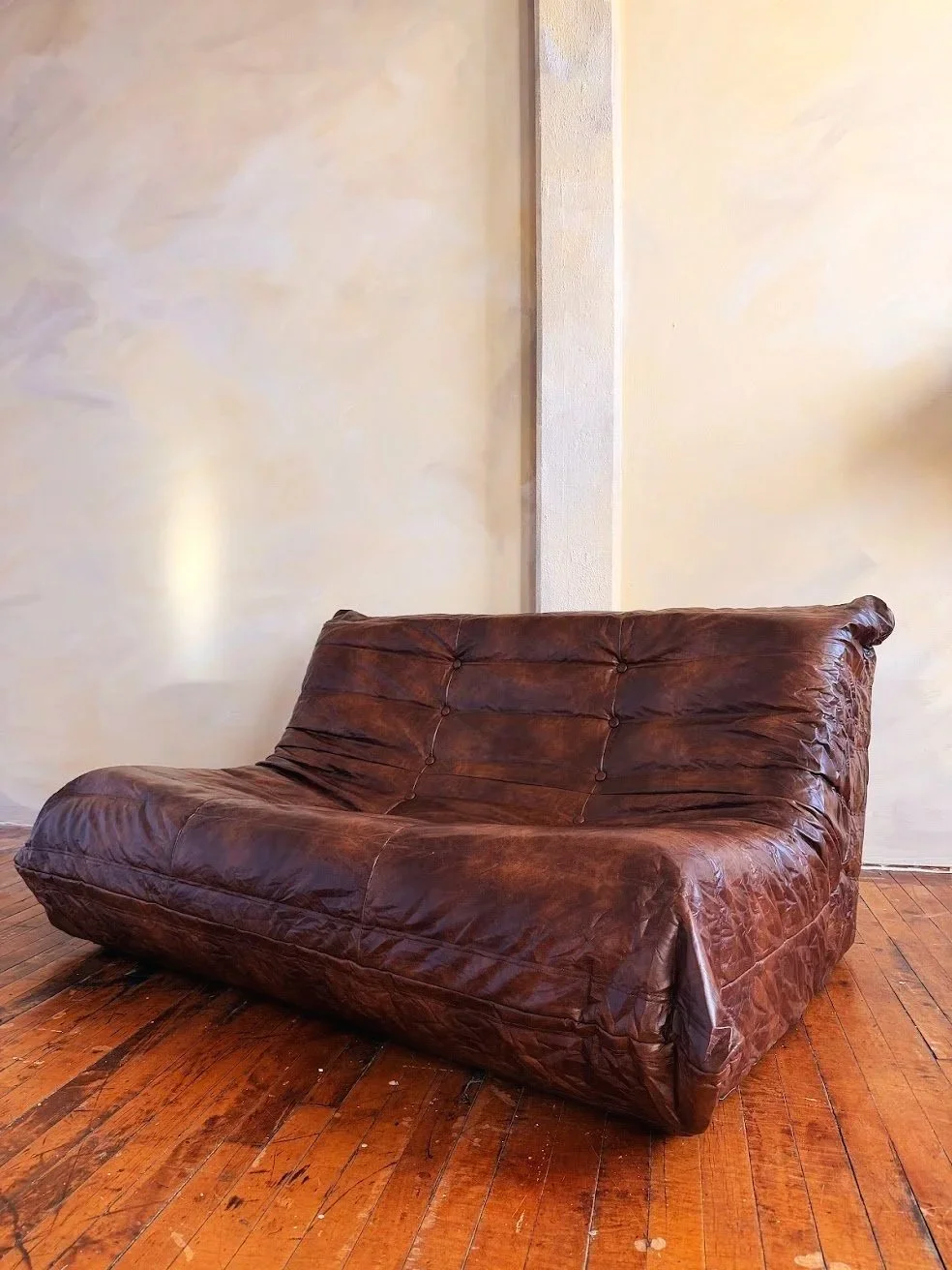 A brown leather couch with a worn appearance, placed on a wooden floor against a beige wall.