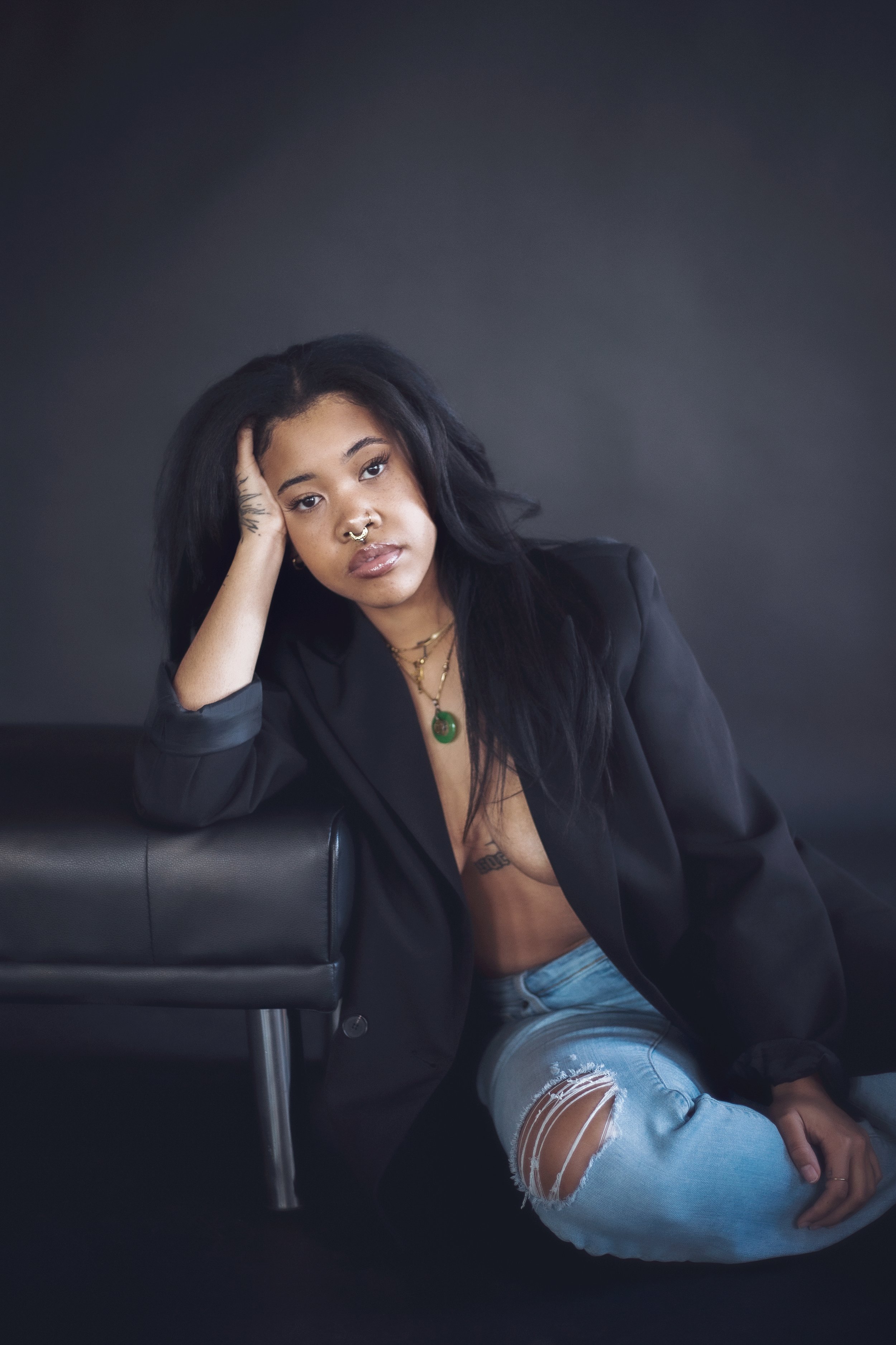Young woman with dark hair, wearing a black blazer and ripped jeans, sitting on the floor against a dark background, with one hand resting on her head and a serious expression.