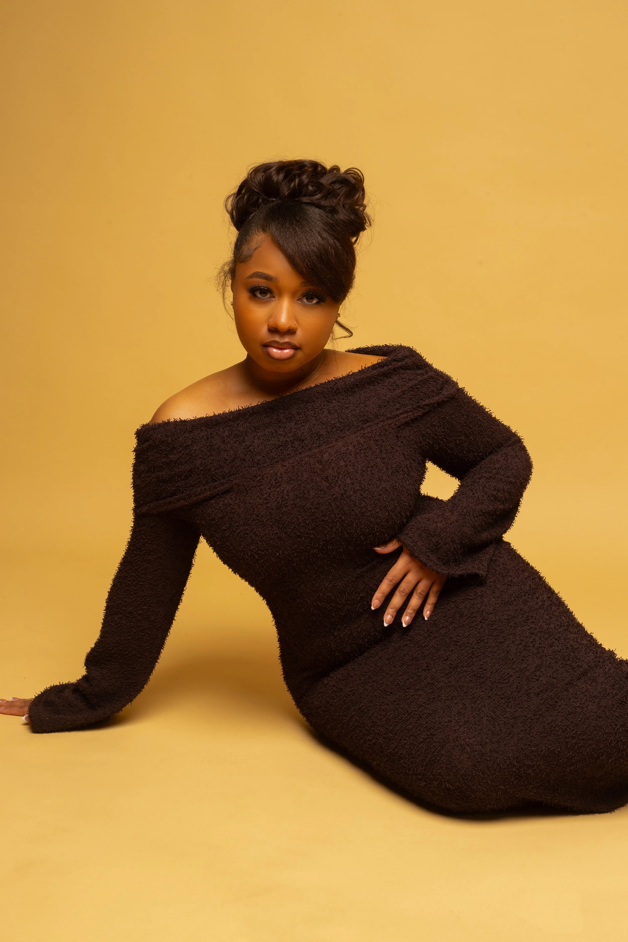 A woman with dark hair styled in large curls and a side-swept bang, wearing a black off-shoulder dress and posing against a yellow background.
